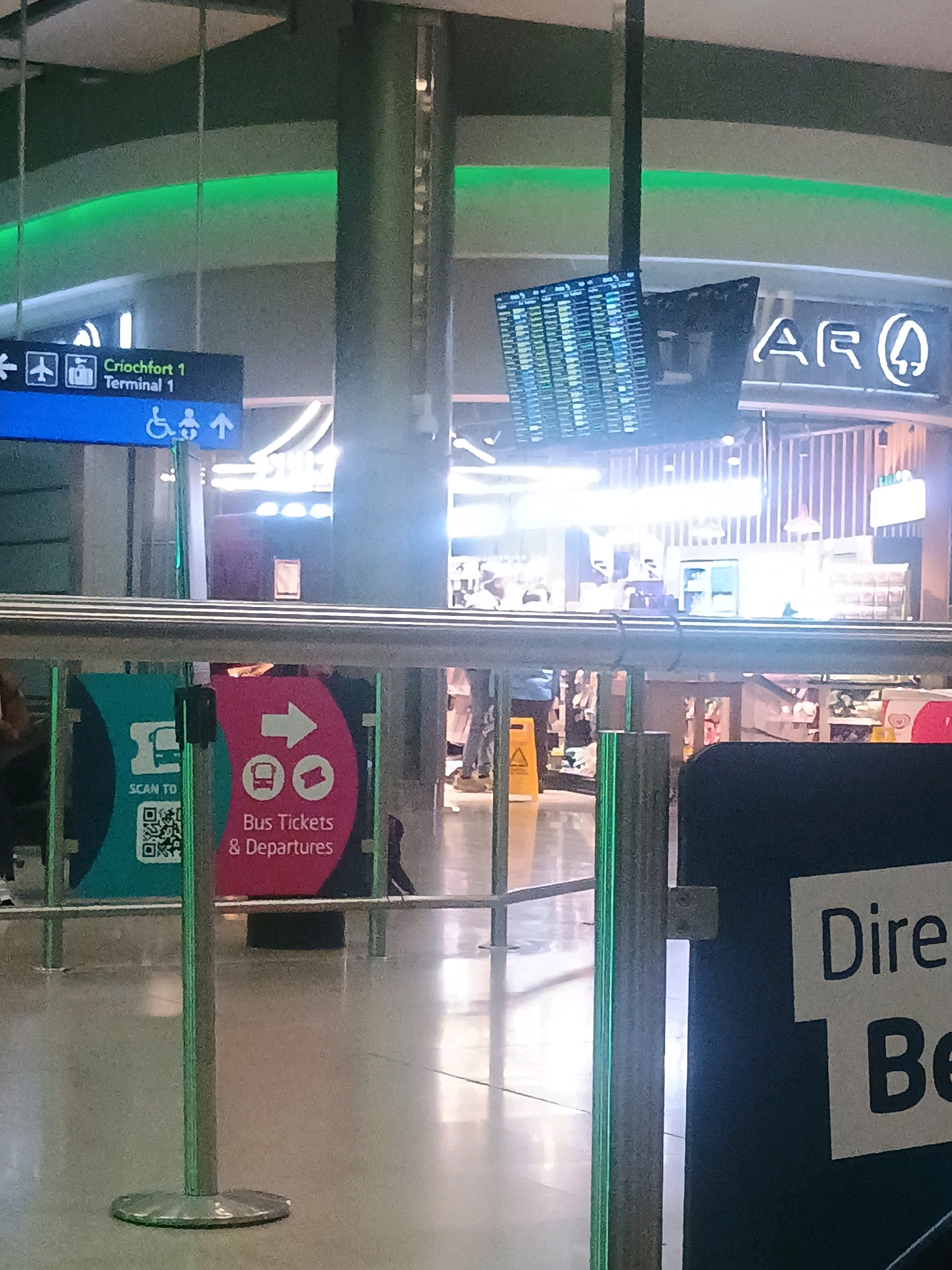 The image shows a sign in what appears to be an airport terminal. The sign is blue and has white text that reads "Criochfort 1 Terminal 1". There are also icons of an airplane, a suitcase, and a wheelchair next to the text. To the right of the sign are icons of a wheelchair and a baby, indicating accessible facilities. In the background, there is a departure board displaying flight information. Further in the background, there is a neon sign with the letters "AR". The floor is tiled and reflective. In the foreground, there is a green stanchion and a black sign with white text that is partially visible and appears to say "Dire Be".