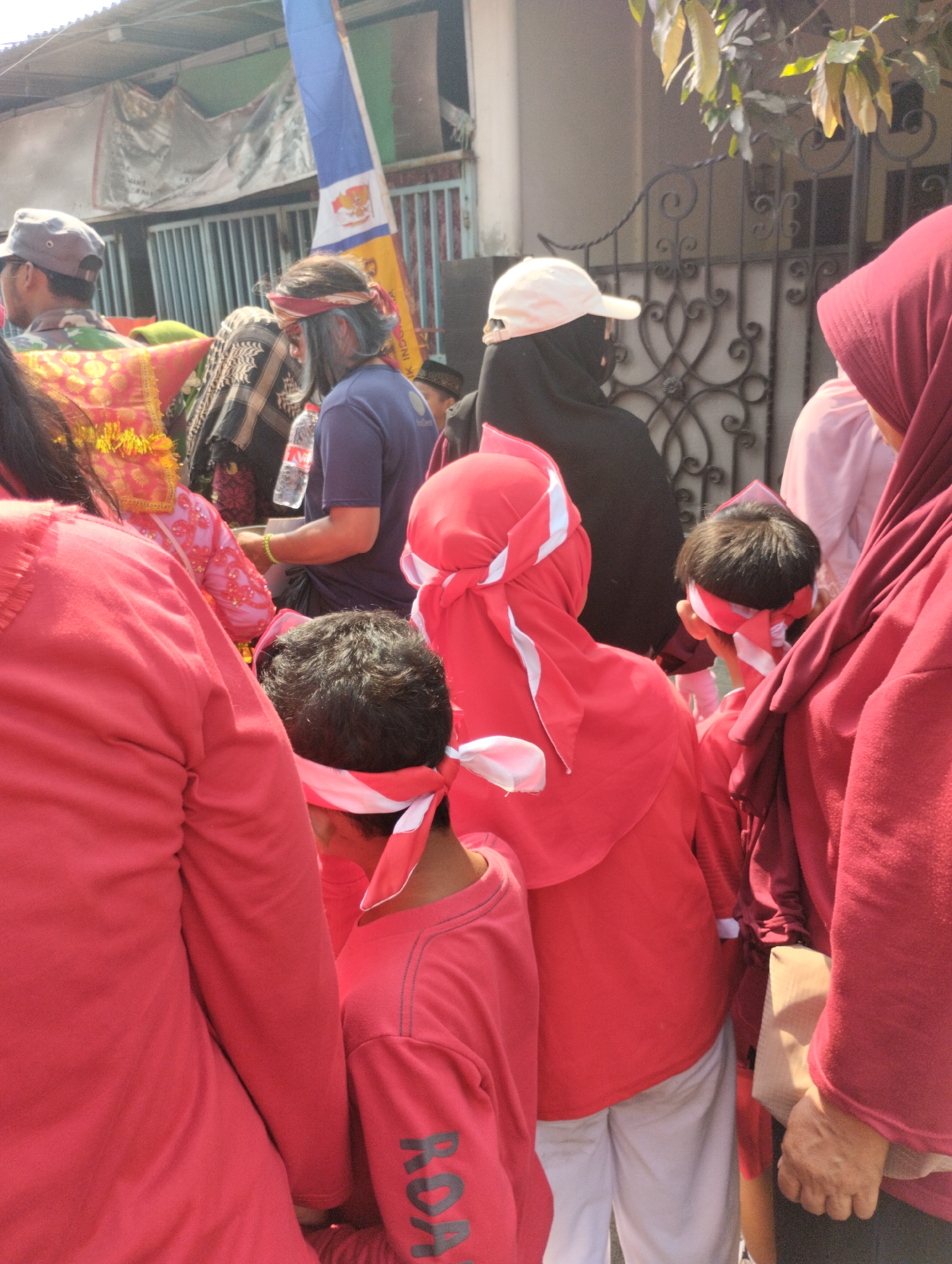 The image depicts a gathering of people, likely participating in an outdoor event or demonstration, in Bekasi, Indonesia. The dominant colors are red and maroon, seen in the clothing of many individuals, including headscarves and shirts. Several people are wearing red headbands with white accents. One individual in the center-left has blue-tinted hair and is wearing a headband. In the background, a flag with white and blue colors is visible, along with buildings and a decorative metal fence. The lighting suggests it is daytime, likely sunny, casting shadows and highlighting the textures of the clothing and environment. The focus is on a group of people from the chest up, with some children among them. The overall impression is of a community event or protest, possibly with a patriotic or cultural theme given the flag.