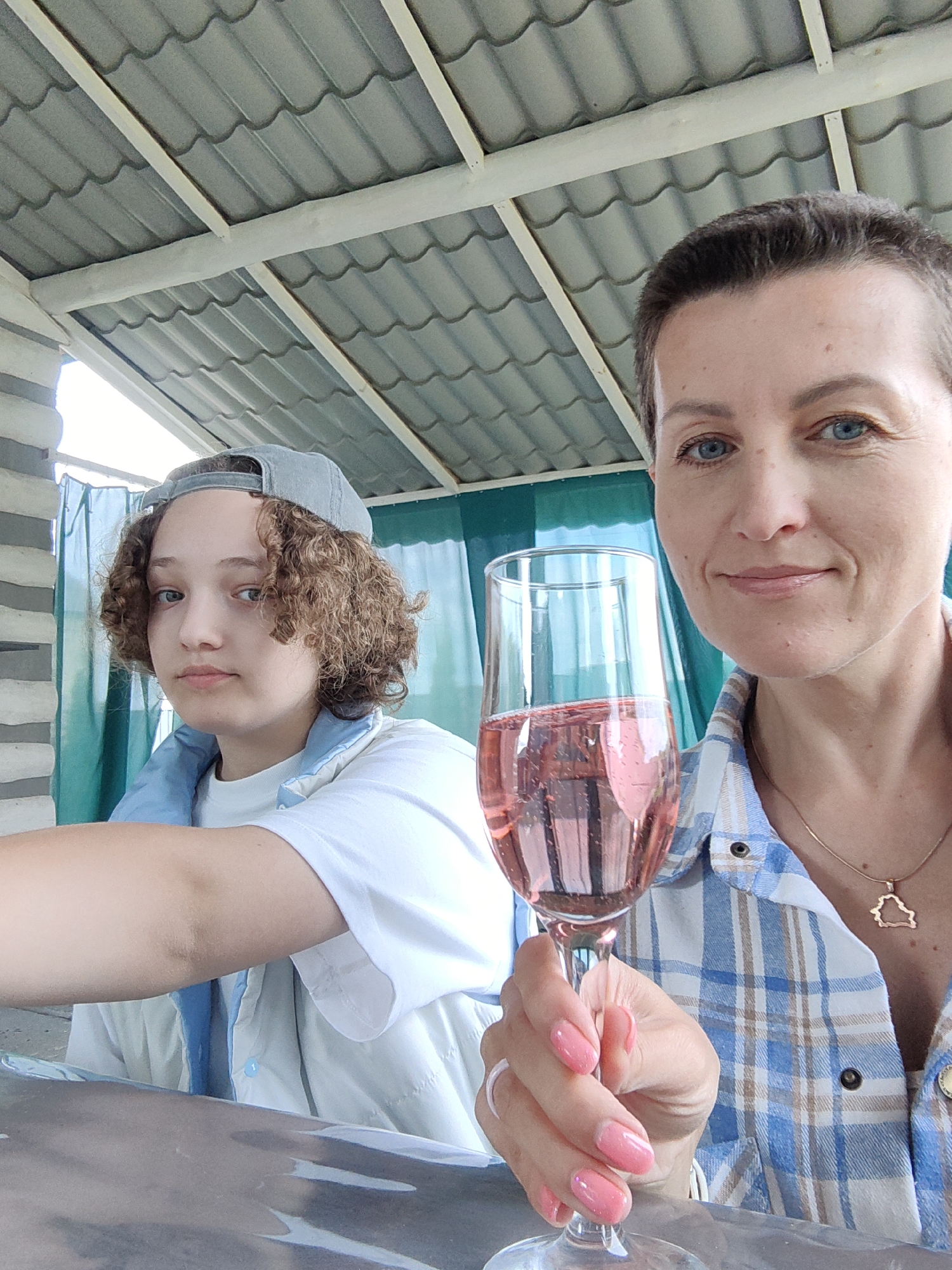 Two individuals are captured in a selfie taken outdoors under a gray, corrugated roof. On the right, a woman with short, dark hair and blue eyes holds a champagne flute filled with pink liquid. She is wearing a blue and white plaid shirt and a gold necklace with a pendant shaped like a simplified map of Belarus. On the left, a younger person with curly brown hair and a gray baseball cap is wearing a white t-shirt and a light blue vest. They are looking towards the camera. The background features a teal curtain and a brick-like pillar. The scene suggests a casual outdoor gathering, possibly in the city of Myadzyel, Belarus, given the pendant's design.