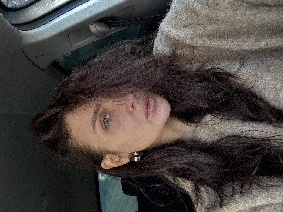An adult female is depicted in a close-up portrait, seated within the interior of a vehicle, in the city of Chaikovsky, Russia. She possesses long, dark brown hair, parted on the side, with a section falling over the right side of her face. Her left eye, green in color, is visible, while her right eye is partially obscured. Her lips are full and appear natural or lightly tinted. A large, reflective, silver-toned hoop earring is present on her visible left ear. She is wearing a light brown or beige knitted sweater with a textured finish. The background reveals the car's dark interior, including a headrest to the left and parts of the A-pillar and roof lining to the right, suggesting the image was captured during daylight hours. The individual is looking directly forward, towards the camera.