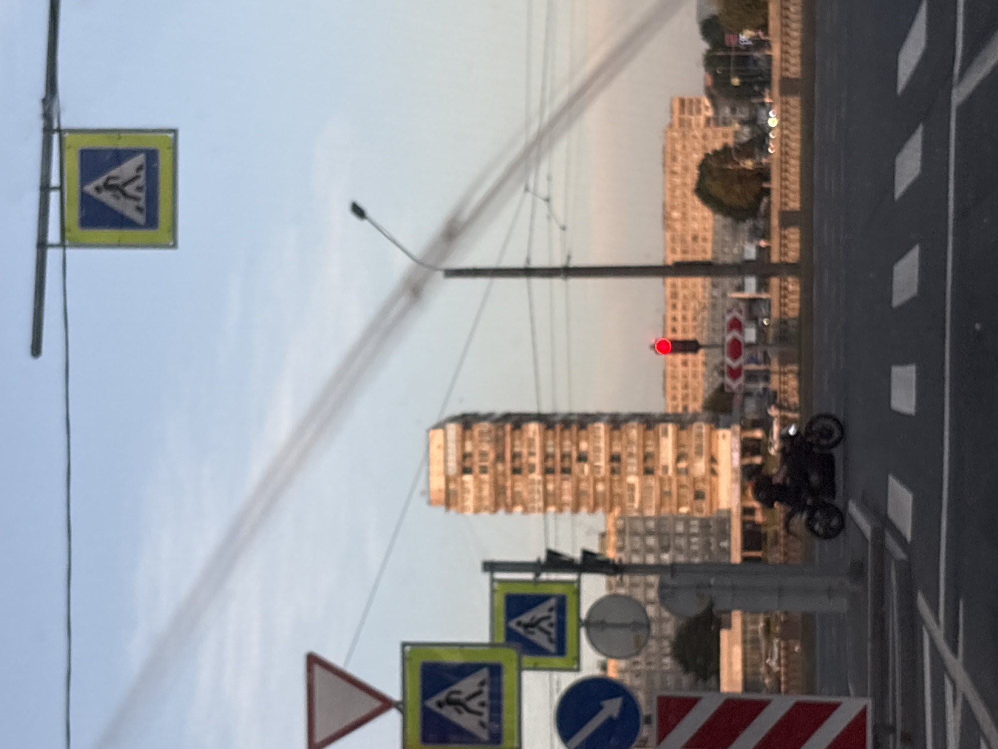 The image shows a street scene in Saint Petersburg, Russia, with a pedestrian crossing in the foreground and a tall residential building in the background. Several traffic signs are visible, including a "Give Way" sign, two "Pedestrian Crossing" signs, and a blue circular sign with a white arrow pointing left. A traffic light is showing red. A person is riding a motorcycle on the street. The sky is light blue with some wispy clouds. The overall lighting suggests it is late afternoon or early evening.