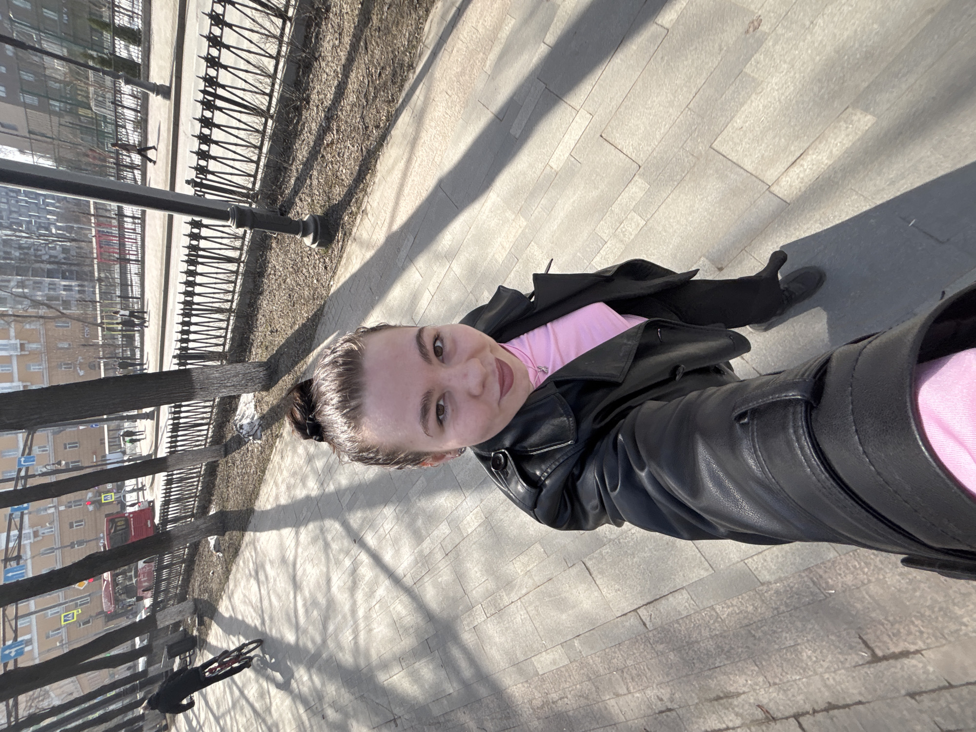 A selfie of a light-skinned person with brown hair, wearing a pink shirt and black leather jacket, is taken on a gray paved walkway, likely in the city of Perm, Russia. A black metal fence with decorative posts runs parallel to the walkway. Buildings and trees are visible in the background. Shadows from the trees fall across the walkway. The person is wearing black gloves and black shoes. An umbrella is also visible along the walkway.