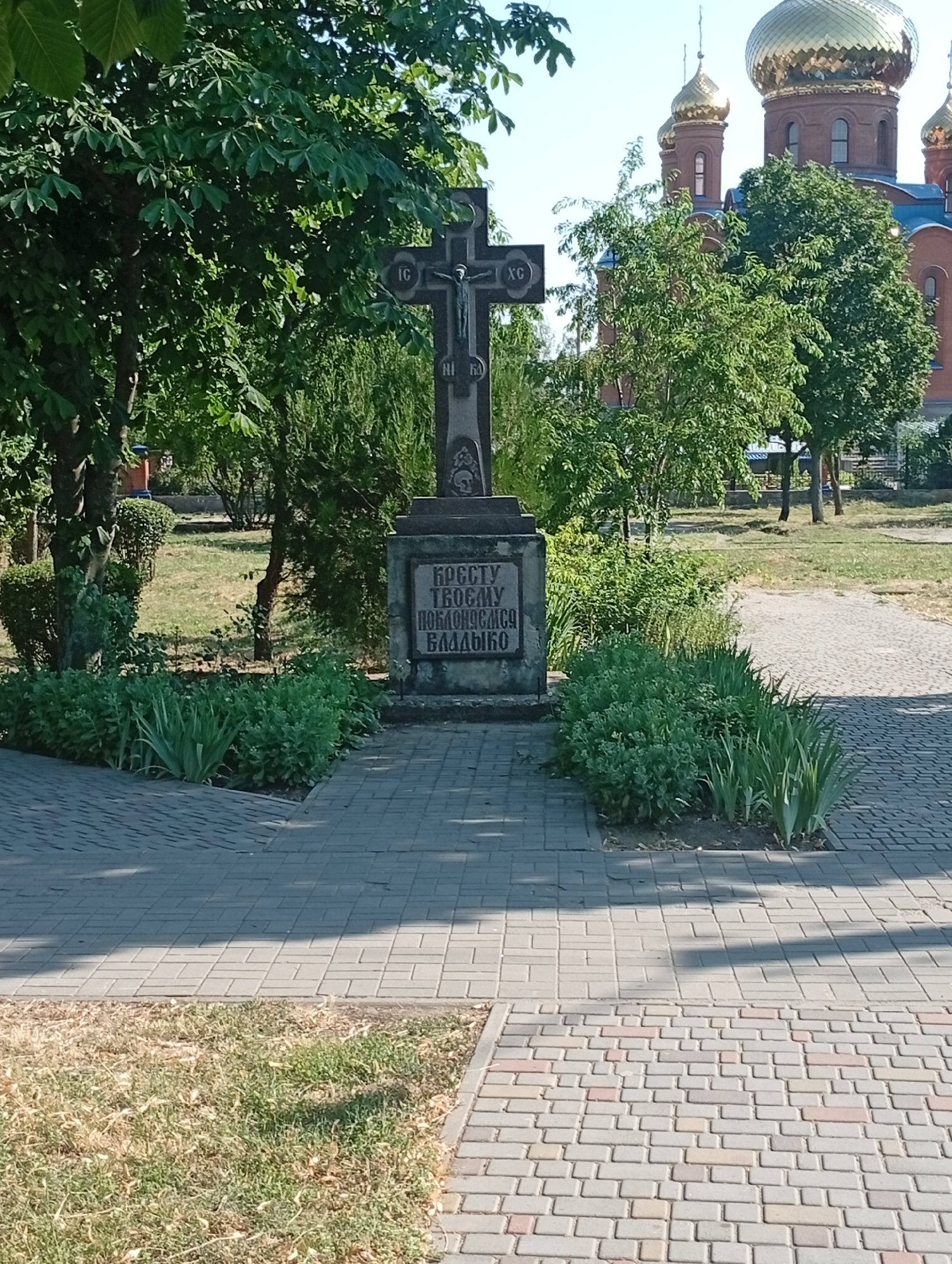 A dark, stone cross stands prominently in the foreground, featuring a depiction of Jesus Christ. The cross is mounted on a stone pedestal with engraved text. Behind the cross, lush green trees and bushes create a vibrant backdrop. In the distance, a large, red church with golden domes is visible, suggesting a religious setting. The scene is bathed in bright sunlight, indicating a clear, sunny day. The ground is paved with cobblestones, and there is a patch of dry, yellowed grass to the left of the path. The overall atmosphere is peaceful and serene. The engraved text on the pedestal appears to read: "КРЕСТУ ТВОЕМУ ПОКЛОНЯЕМСЯ ВЛАДЫКО". The image is captured from a slightly low angle, looking up towards the cross and the church. The time of day is likely daytime, given the strong sunlight and clear sky. The location is identified as Pokrov, Ukraine.