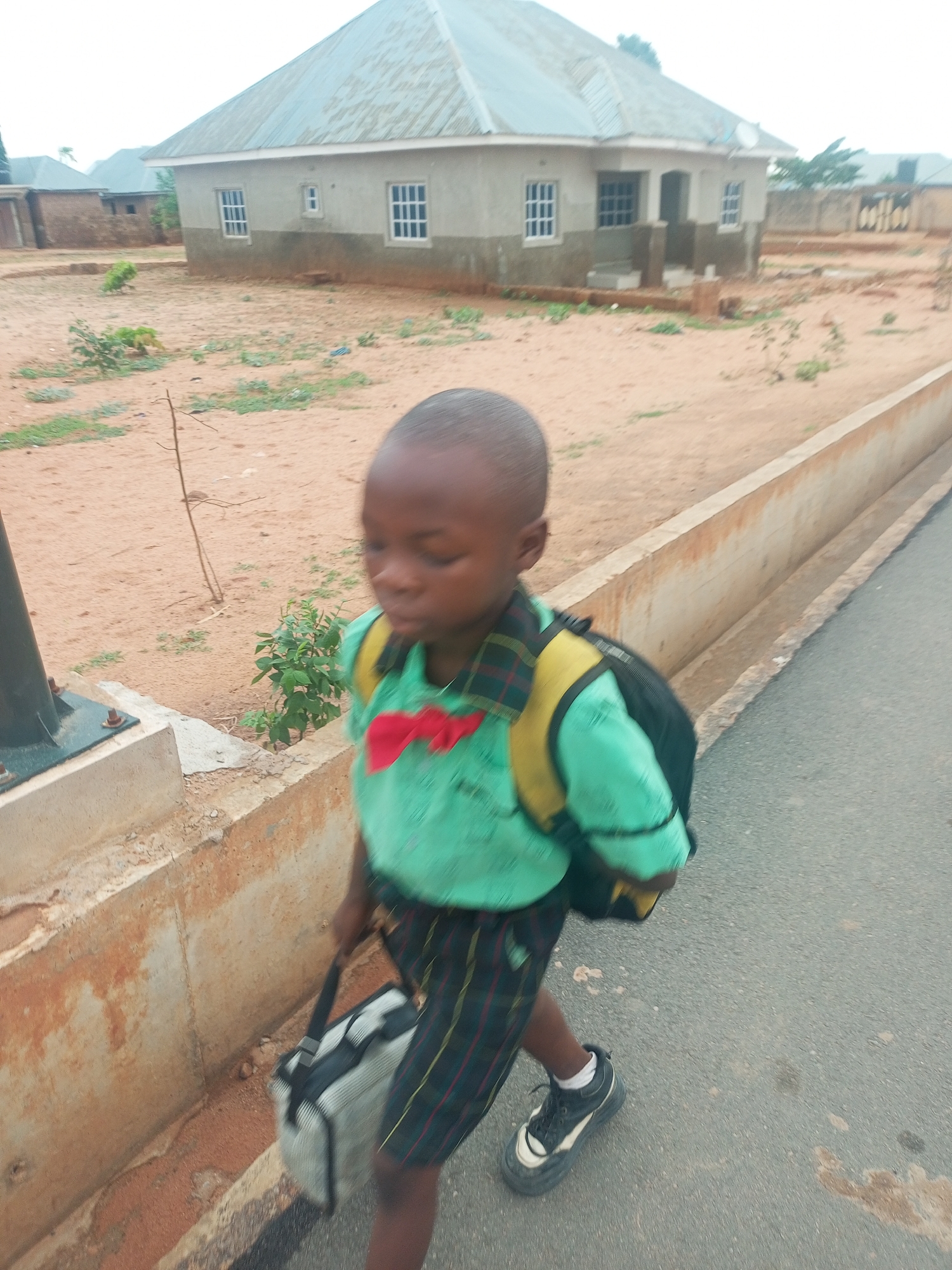 A young boy is walking along a paved road. He is wearing a green shirt with a red bow tie, plaid shorts, and sneakers. He has a backpack on his back and is carrying a gray bag in his hand. To his right is a low concrete wall. Behind him are houses in a neighborhood with dirt yards. The image was taken in the city of Tempure, Nigeria.