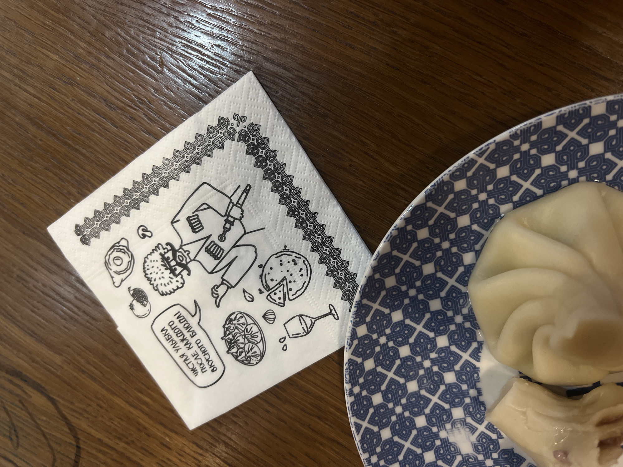The image shows a white napkin with black printed illustrations and text, placed next to a plate with food, on a wooden table. The napkin features a cartoon-like figure with a chokha (traditional Georgian coat), along with sketches of different food items, and a text bubble in Russian. The plate has a blue and white geometric pattern and contains khinkali, a type of Georgian dumpling. The lighting suggests an indoor setting, possibly in a restaurant or cafe. Although it is hard to be certain, the text on the napkin implies that this picture may have been taken in Georgia. It is impossible to determine the location in the city of Izhevsk, Russia.