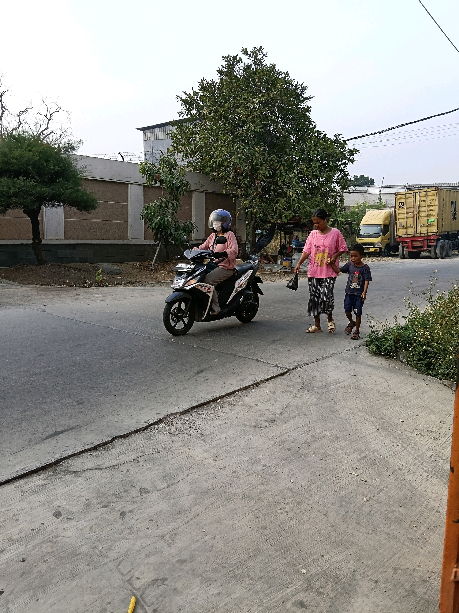 A woman wearing a pink shirt and black and white striped skirt, along with a young boy in a dark blue shirt and shorts, are walking side-by-side on a paved road. The woman is holding a black plastic bag in her left hand and her right arm is around the boy's shoulders. The boy is looking forward and walking slightly ahead of her.
In the background, a woman wearing a light pink long-sleeved shirt and grey pants is riding a black motorcycle. She has a blue helmet on and a white mask covering her face. The motorcycle has a license plate with the text "B 4675 JIT".
Further in the background, a yellow truck with a shipping container is parked on the side of the road. The container has text that is partially visible, including "MCM 114767 9" and "MSC". Behind the truck and container, there are buildings and trees. The scene appears to be an outdoor setting with a clear sky, suggesting it is daytime. The road is paved with concrete, and there is a line of green bushes on the right side of the image. The overall setting suggests an urban or industrial area in Belimbing, Indonesia.