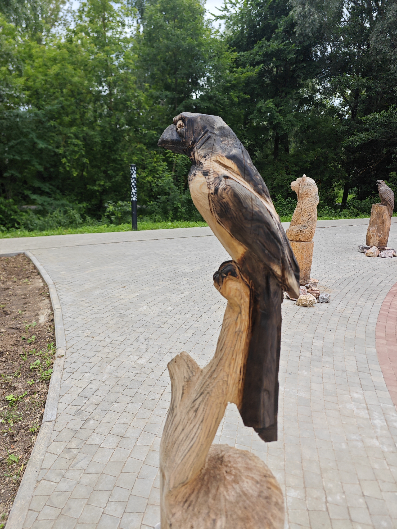 A wooden sculpture of a crow, carved from a log, is positioned in the foreground of this outdoor scene. The crow sculpture is dark brown and black, with lighter wood visible on its chest and wings. It perches on a rough-hewn wooden branch that extends from a larger log base.
Behind the crow sculpture, a paved walkway made of interlocking grey diamond-shaped pavers curves into the background. To the right of the walkway, there are other wooden sculptures: a bear standing upright and a bird of prey perched on a log. These sculptures are also made from natural wood, exhibiting varied grain patterns and colors. The ground around the sculptures is covered with small stones.
In the background, a lush green forest lines the edge of the walkway. The sky above is overcast, with patches of lighter sky visible through the leaves. A black lamppost with white dotted patterns stands in the middle ground, near the edge of the forest. The overall setting appears to be a park or outdoor recreational area in Yoshkar-Ola, Russia. There are no people visible in the image.