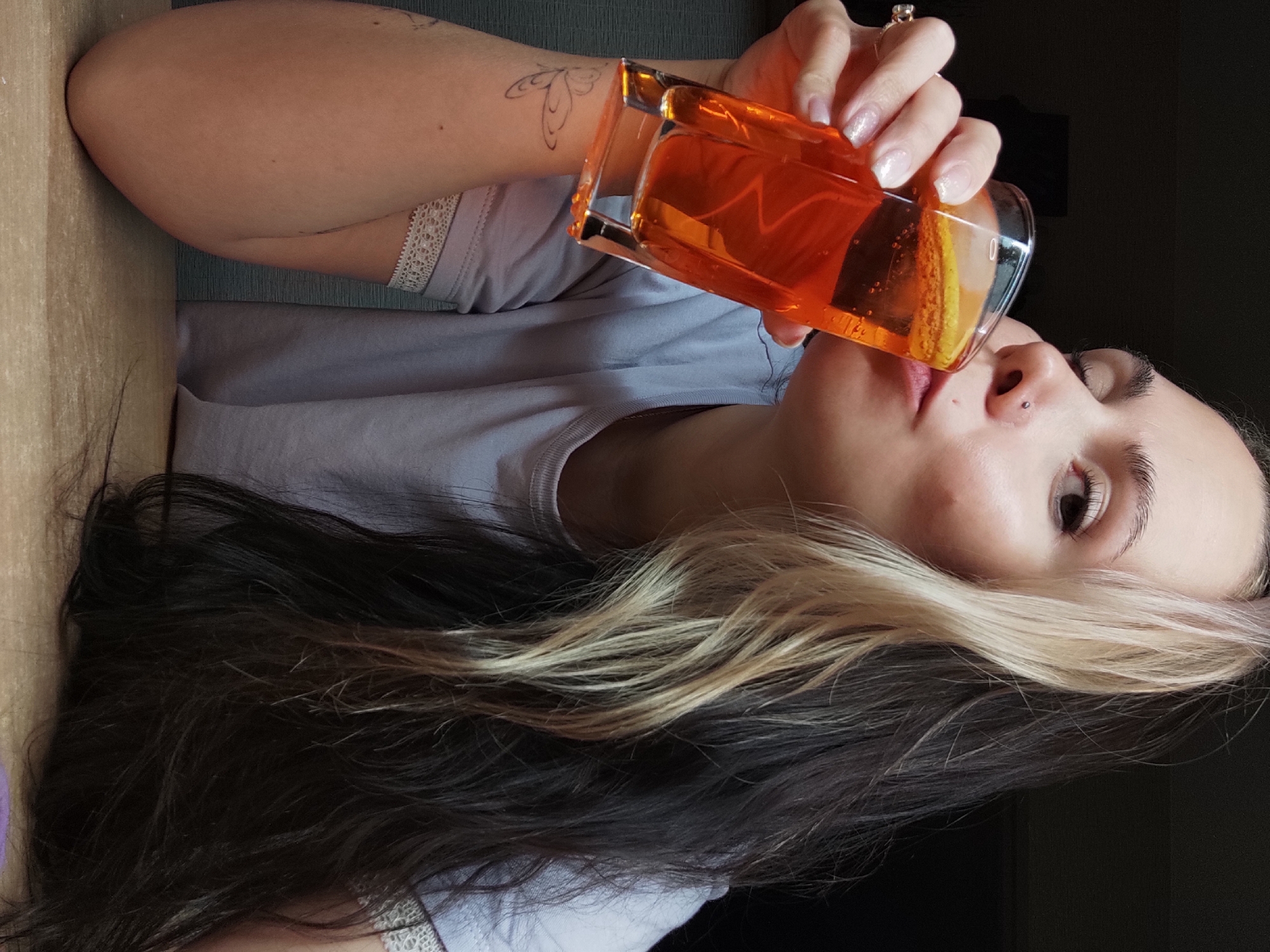 A woman with dark hair with a blonde streak is drinking from a rectangular glass containing an orange liquid. She has fair skin and a nose piercing. She is wearing a light gray t-shirt. The setting appears to be indoors, with a wooden surface to the left of the woman. A tattoo is visible on her left arm. The exact location within the city of Chaikovsky, Russia, is not discernible from the image.