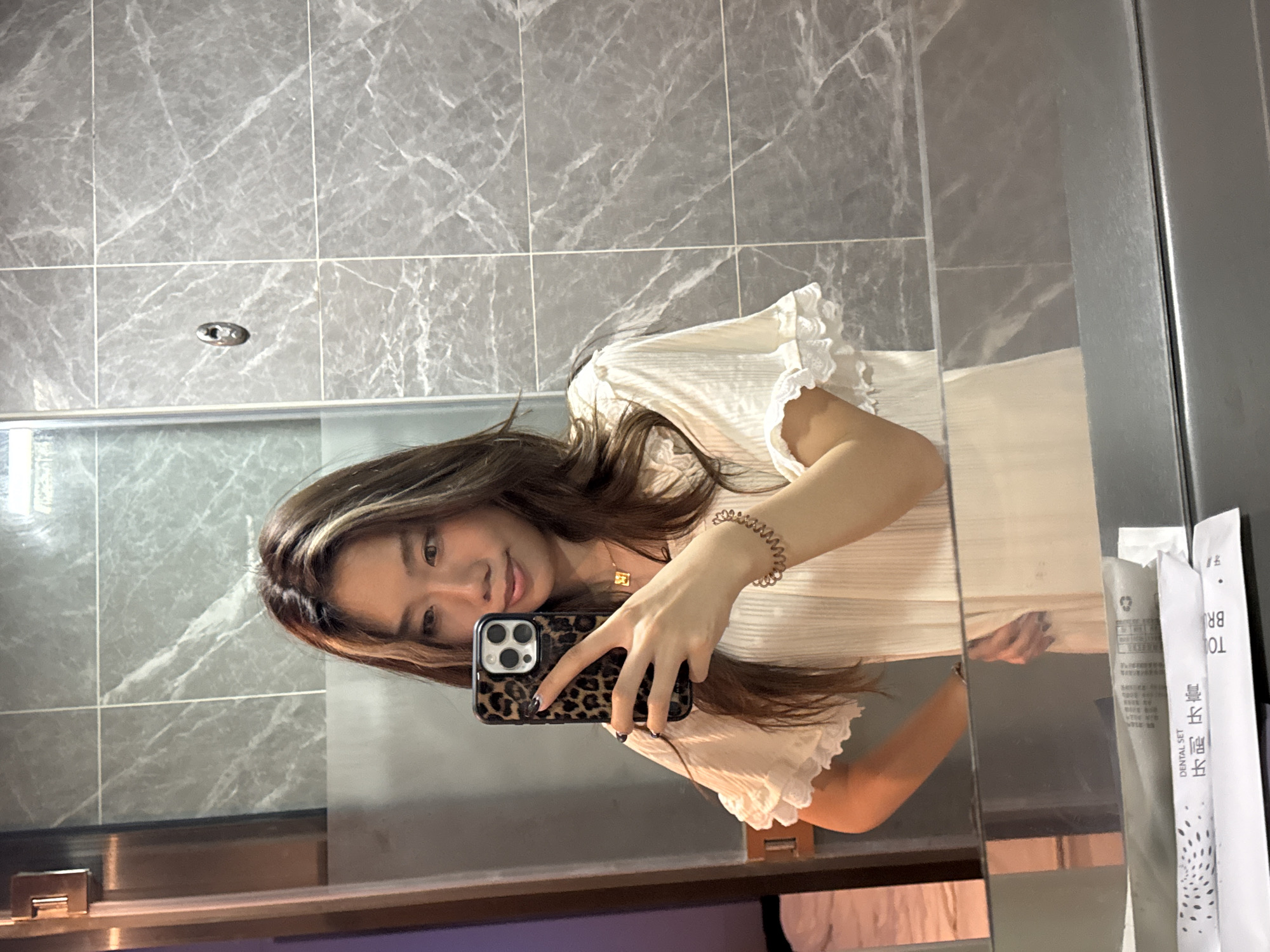 A woman with light brown hair with darker streaks is taking a selfie in a bathroom. She is holding a phone with a leopard print case in her left hand. The bathroom has grey marble-like tiles on the wall. On the right side of the image, there are toothbrush sets. The location is Chenghua District, China.