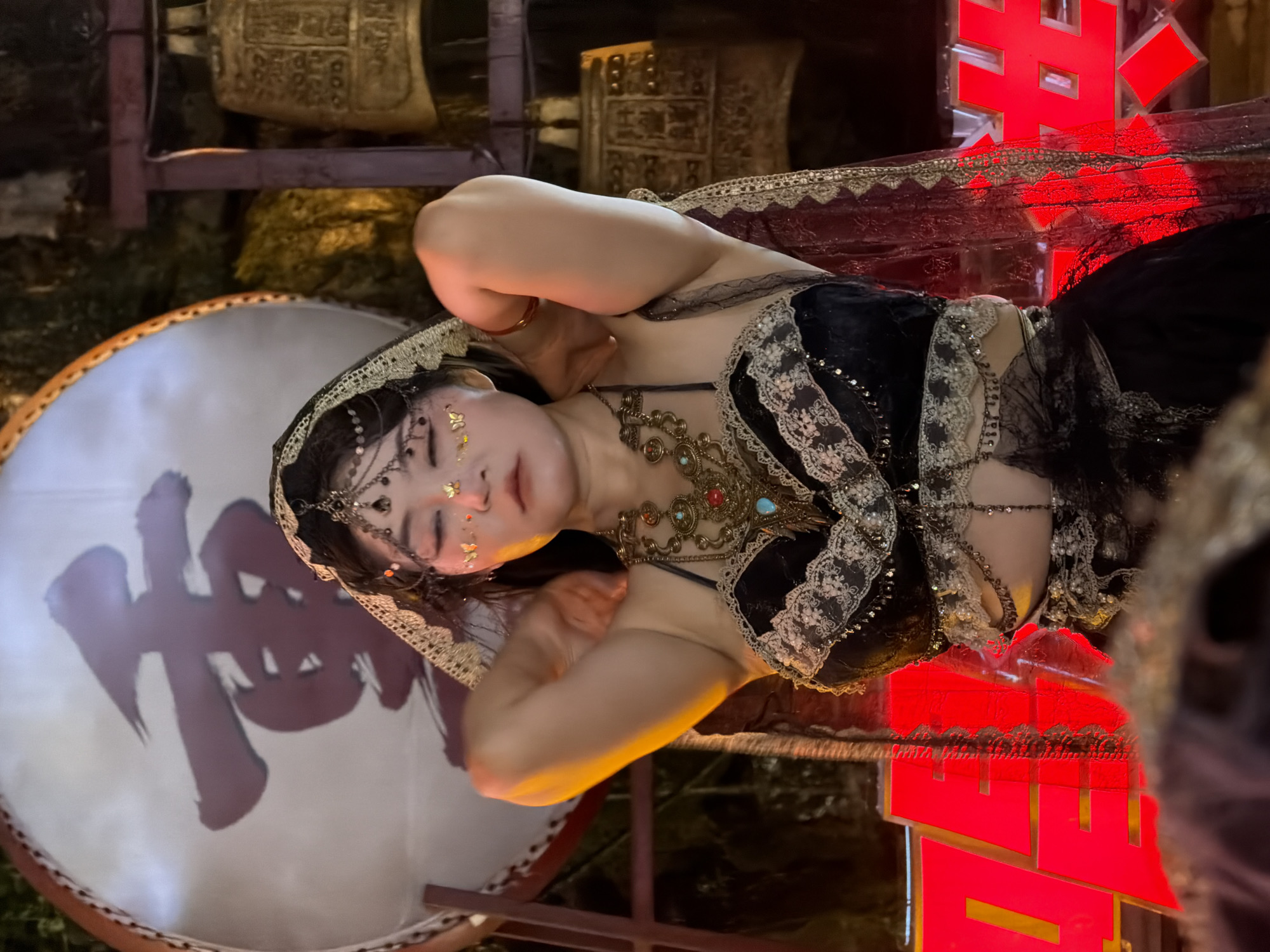 In Yuzhong District, China, a woman is pictured at night, dressed in a black belly-dancing costume adorned with lace and beads. She wears a headpiece with dangling ornaments and has decorative gold glitter on her face, including butterfly shapes near her eyes. She is adjusting her costume, with her arms raised and hands near her hair. In the background, a large drum with a red Chinese character is visible, along with several large brass bells suspended from a wooden frame. Red neon lights glow at the bottom of the frame, suggesting an urban setting. The overall scene has a festive or performance-oriented atmosphere.
