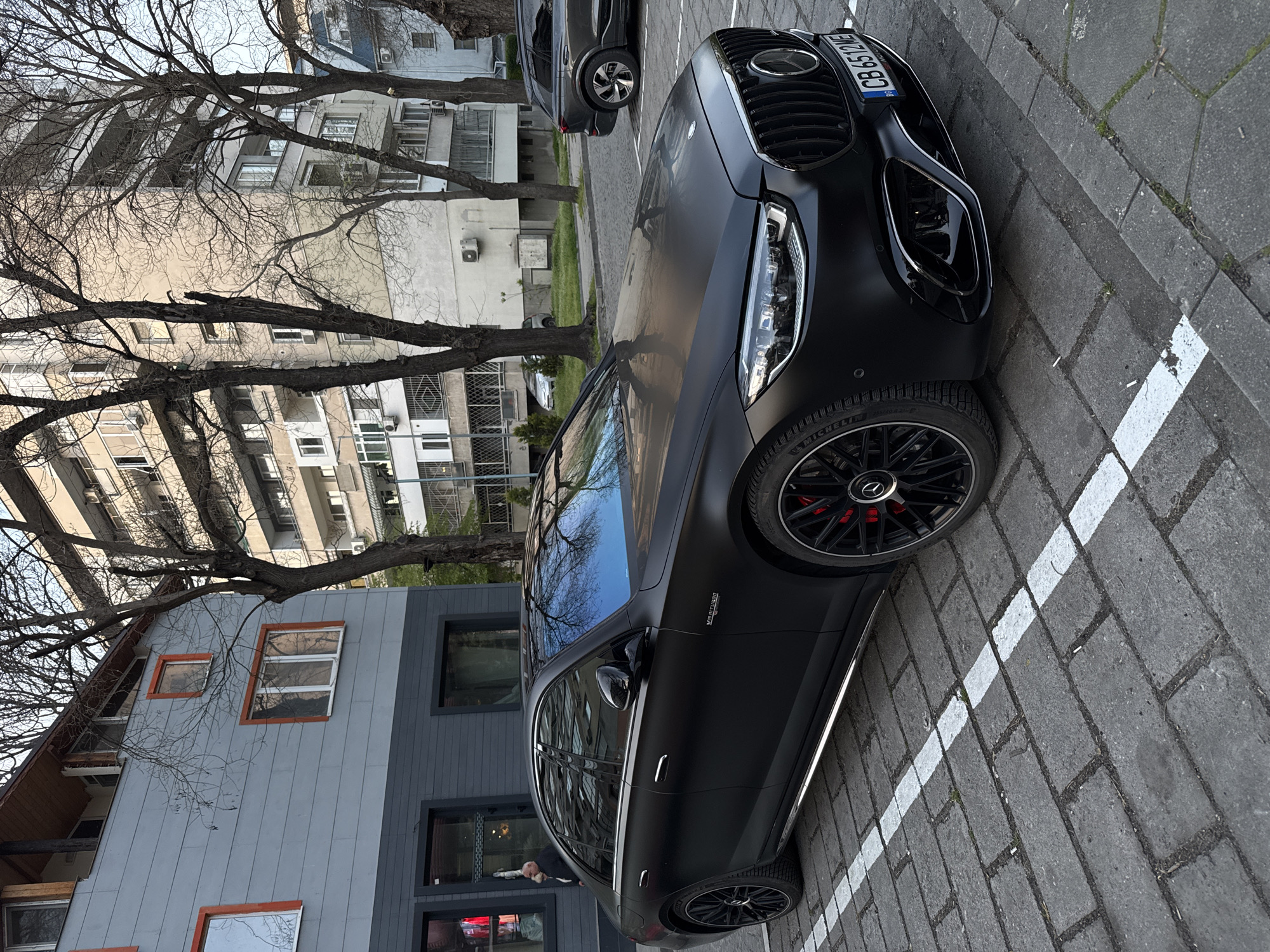 A black Mercedes-Benz AMG sedan is parked in a designated parking spot on a brick-paved street in an urban setting. The car is angled slightly towards the camera. The front of the car, including the grille and headlight, are visible. The license plate number is CB 6512 HE. The car has black rims with red brake calipers. The building directly behind the car is two-toned gray, with what appears to be a commercial storefront on the ground floor. Another car is partially visible parked further up the street. Trees with bare branches are also visible. Older residential buildings are in the background. The location is likely in the city of Plovdiv, Bulgaria.