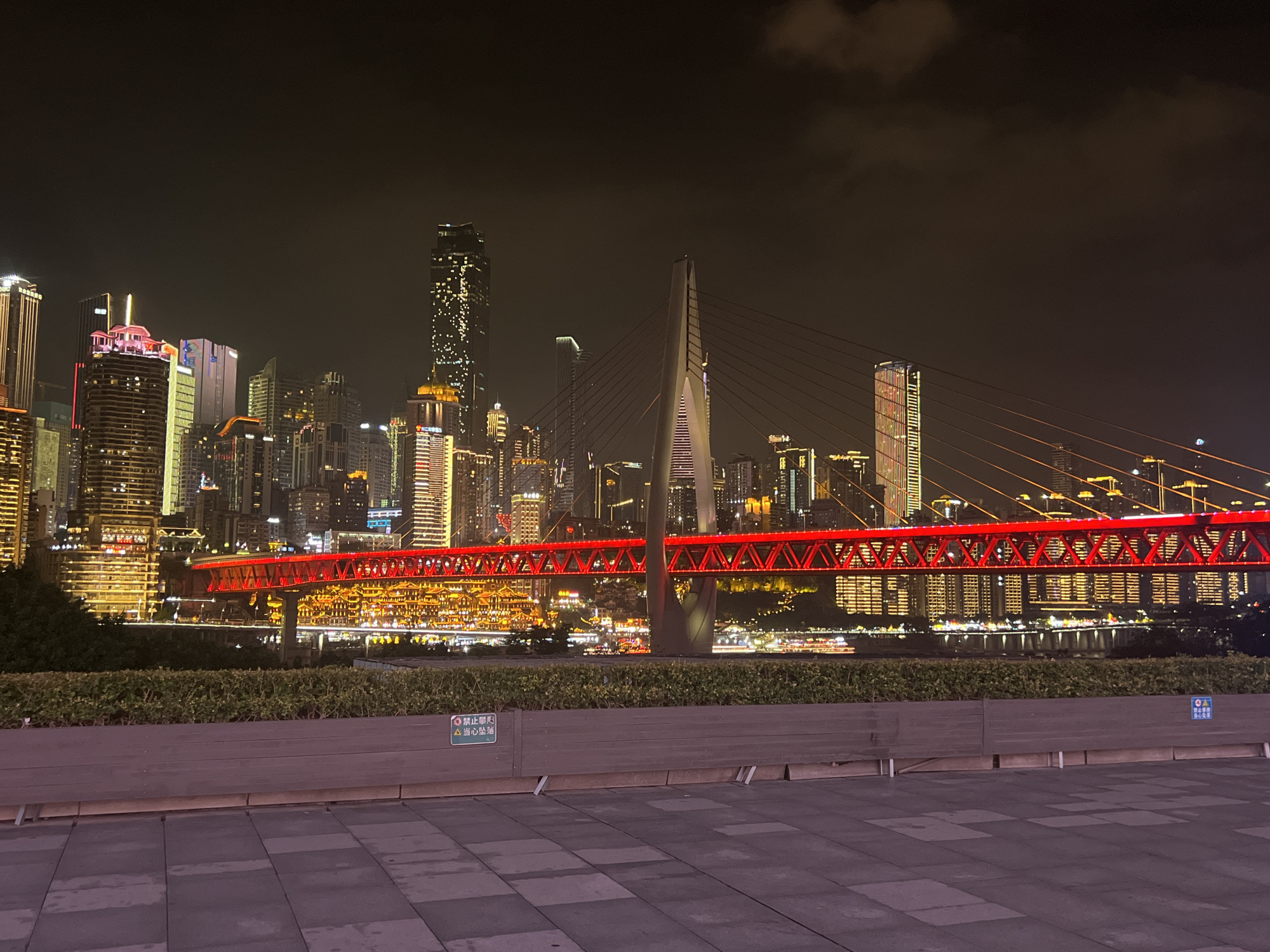 The image shows a nighttime view of the Jiangbeicheng Street area in China, focusing on a brightly lit suspension bridge spanning a body of water. The bridge itself is a prominent feature, with its red illuminated structure and supporting cables stretching across the frame. In the background, a cityscape is visible, characterized by numerous tall buildings adorned with various colored lights, indicating a bustling urban environment. The sky is dark, with some clouds present. In the foreground, there is a paved area with a planter box containing green foliage. A sign with Chinese characters is affixed to the planter box. The scene is illuminated by the artificial lights of the city and the bridge, creating a vibrant and modern atmosphere.