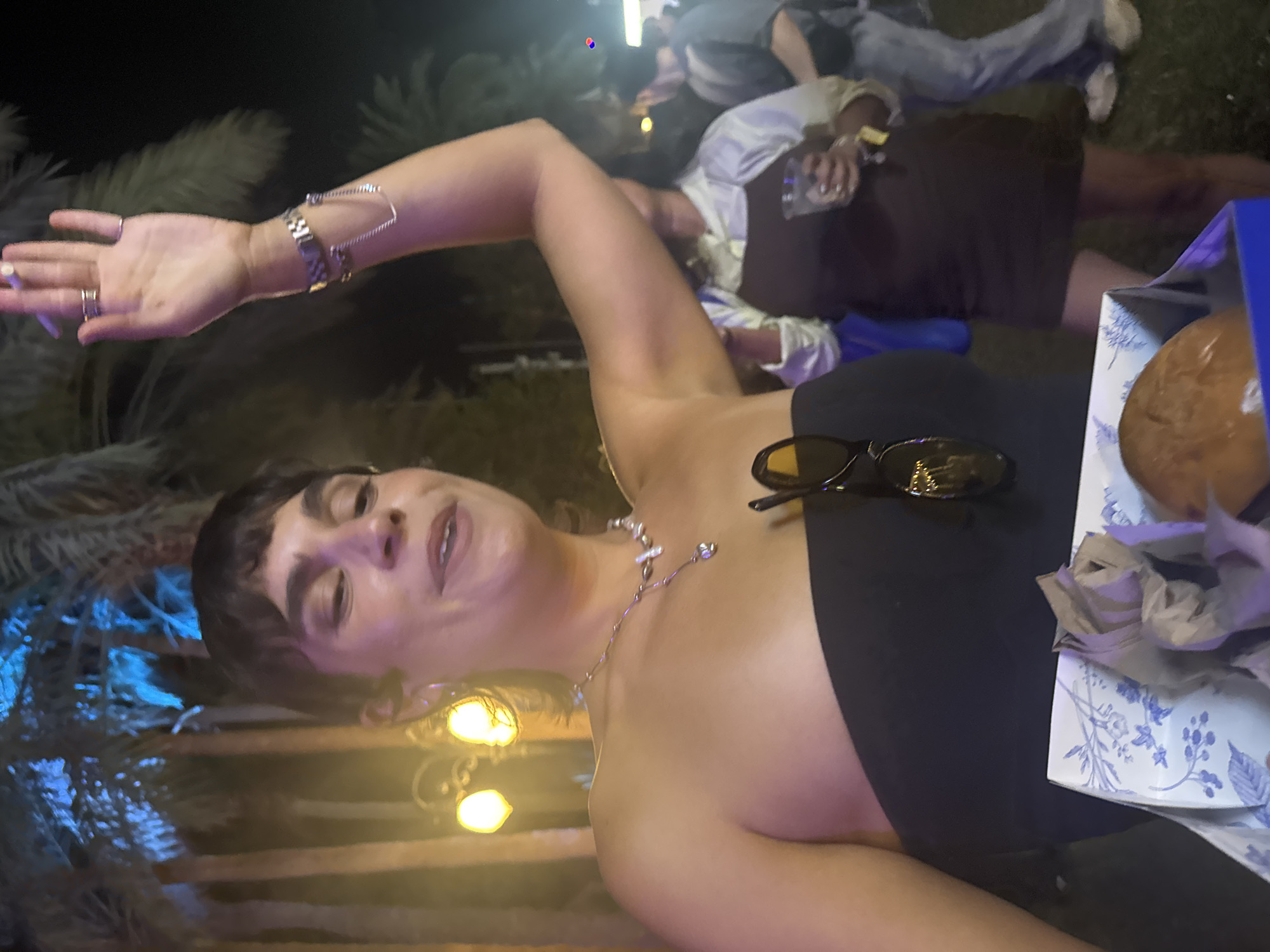 The image shows a woman with short dark hair and a black strapless top, wearing sunglasses and jewelry, with her arm raised and holding a cigarette. A plate with food and a partly visible blue box are next to her. The scene appears to be outdoors at night, possibly in La Quinta, United States of America, based on the available context, with palm trees and a decorative light fixture visible in the background. Other people are in the blurred background.