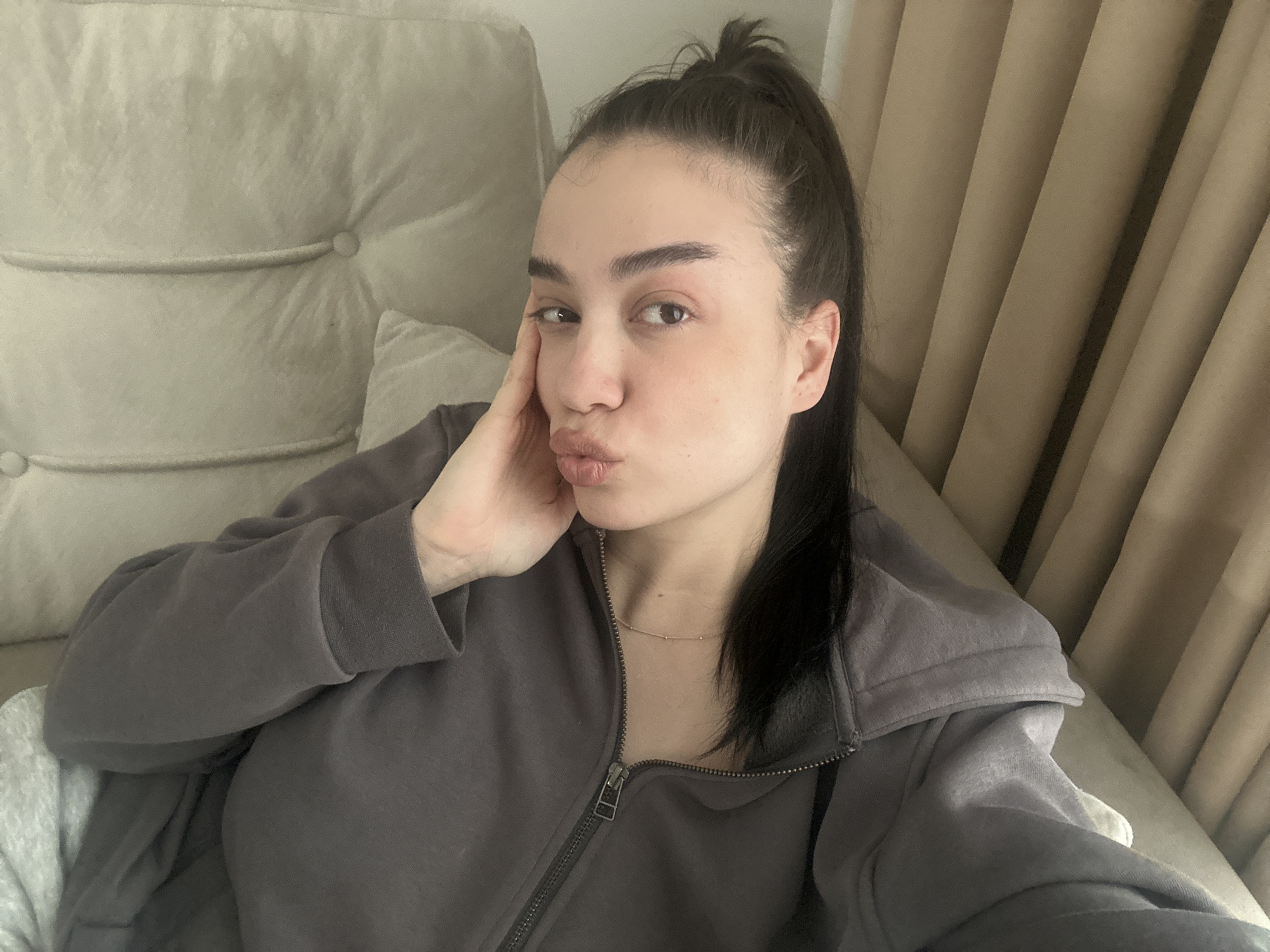 A woman is seated on a beige tufted sofa, wearing a zippered grey hoodie and a delicate necklace. Her dark hair is pulled back into a high ponytail, and she is pursing her lips in a pouting expression while resting her left cheek on her hand. Behind the sofa are tan curtains. There is no indication of this photo being taken in Sofia, Bulgaria.
