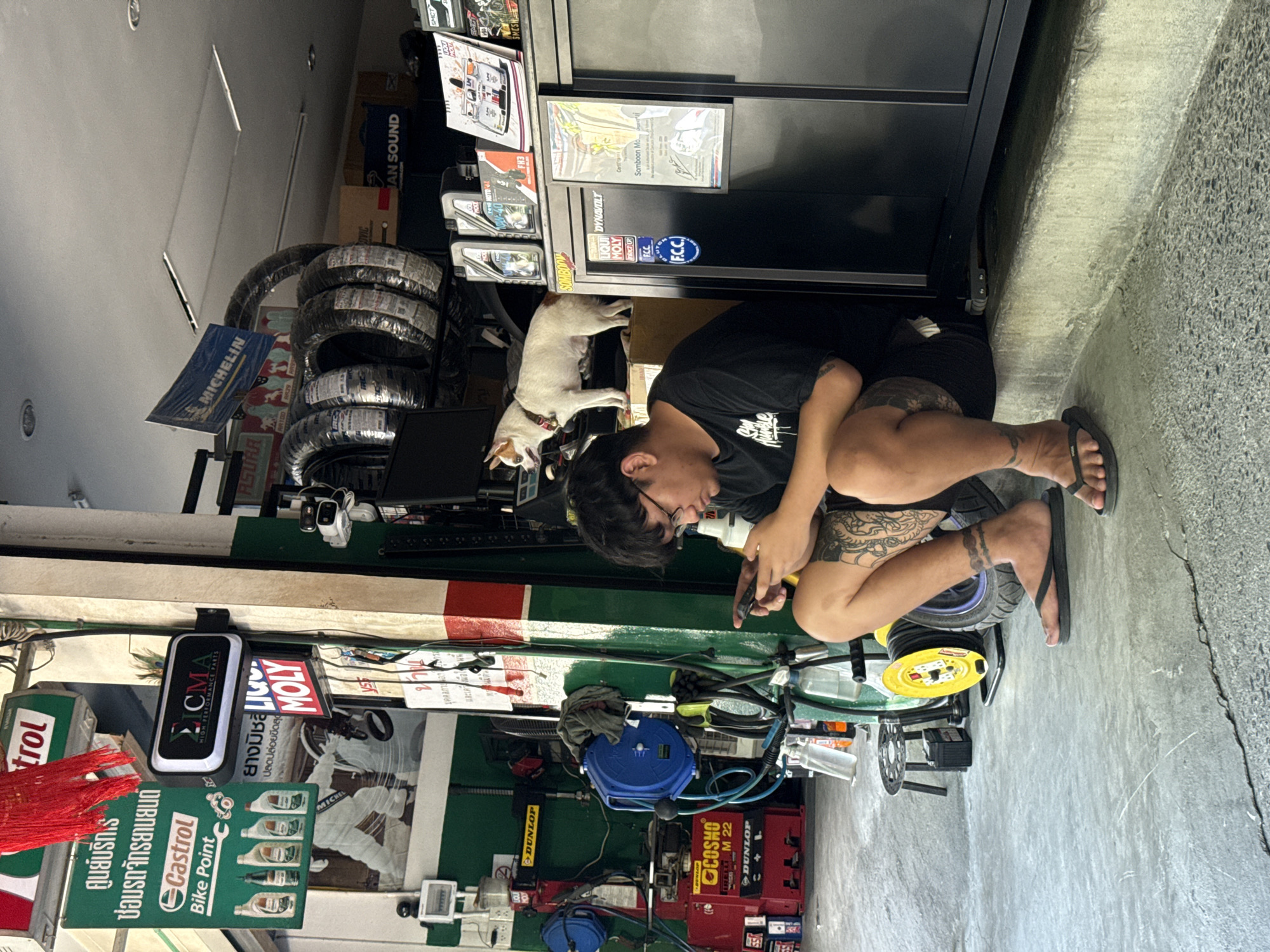 The image depicts an outdoor scene at a motorcycle repair shop in the Ko Samui District, Thailand. A young man with tattoos on his legs and arms is seated on a concrete curb, looking down at his smartphone. He is wearing a black t-shirt and shorts, and flip-flops. Beside him, a small white and brown dog with a red collar stands on top of a cabinet, facing left. The shop's interior, visible in the background, contains various motorcycle parts and equipment, including stacks of tires wrapped in plastic, shelves stocked with oil bottles and other products, and a tire-changing machine. Signs in Thai and English are displayed on the walls and pillars. The environment appears to be a typical roadside workshop with concrete flooring and a corrugated metal roof structure. The lighting suggests it is daytime, possibly late morning or early afternoon.