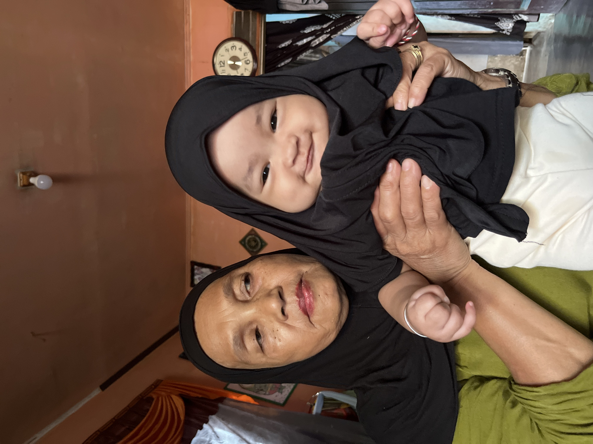 In a medium shot, an elderly woman wearing a black hijab holds a baby also wearing a black hijab in her arms. The woman is looking off to her left, with a slight smile. The baby is looking at the camera with a wide smile. Both are facing forward. The woman's left arm is visible, supporting the baby. The baby is wearing white pants. In the background, a clock is visible on the wall, a light bulb hangs from the ceiling, and draped curtains can be seen. The setting appears to be indoors, possibly a residential interior in Kruengmate, Indonesia.