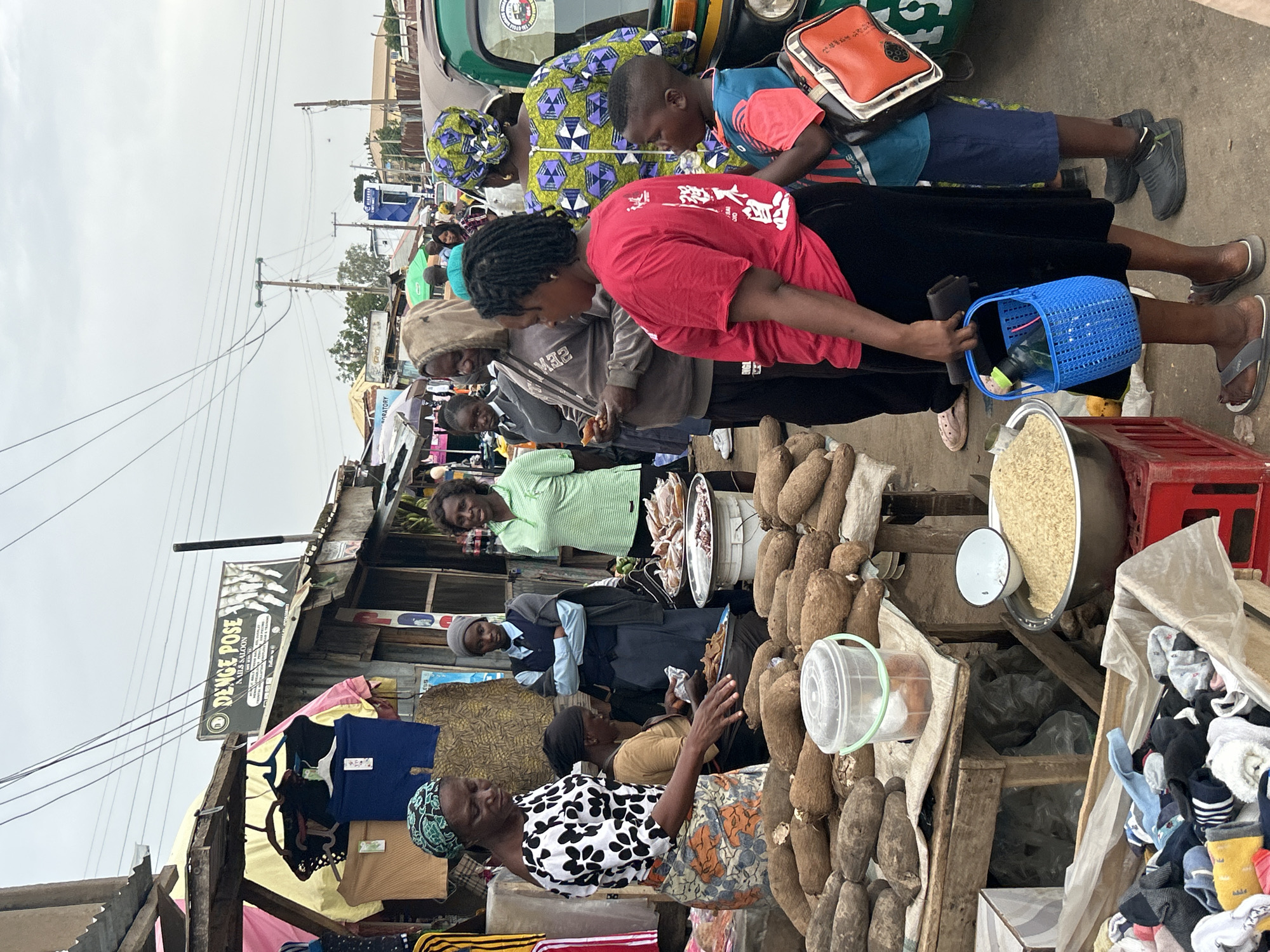 This image depicts a vibrant, busy outdoor market scene in Jos, Nigeria, under an overcast sky. Numerous people, including shoppers and vendors, are present, engaging in daily commerce.
In the foreground and midground, a woman wearing a black and white patterned top and a colorful wrapper stands near a wooden stall laden with a large quantity of root vegetables, likely yams. Adjacent to these, a large metal bowl filled with a light-colored grain, possibly rice, is displayed next to a red plastic crate.
Central to the image, a woman in a red t-shirt with white Chinese characters on the back holds a blue plastic basket containing a bottle, appearing to be a customer examining goods. Beside her, a child in a blue and red shirt with dark shorts carries a brown satchel while standing next to a woman in a patterned traditional outfit and a green vehicle, likely a 'keke Napep' (auto-rickshaw). A woman in a grey hoodie is also part of this group.
Other individuals are scattered throughout the market. A woman in a light green striped shirt stands behind a stall displaying what appears to be prepared food or snacks. Market stalls are constructed from wood and corrugated metal. Various clothing items, including t-shirts and socks, are visible on display.
Overhead, power lines run across the frame, connecting to utility poles, suggesting an urban or semi-urban environment. A prominent black and white sign on the left side reads "D'ENGE POSE NAILS SALOON," indicating nearby services. The scene conveys a lively and bustling market atmosphere, typical of a local Nigerian marketplace.