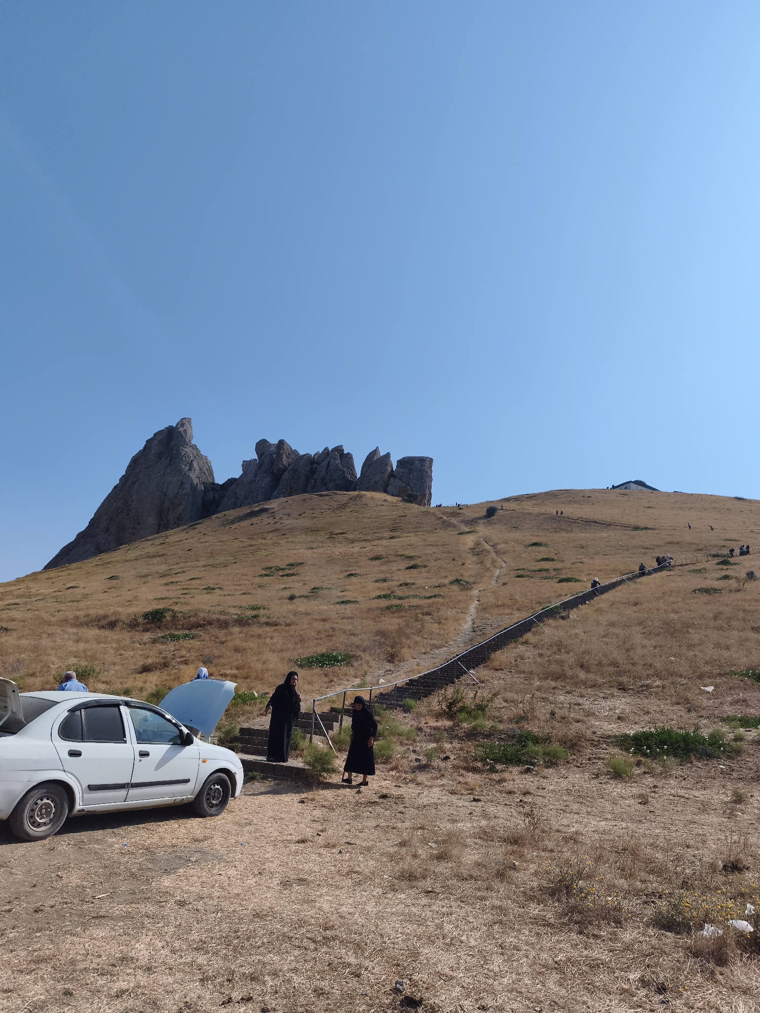 An outdoor, low-angle, eye-level shot depicts a group of people near a white sedan parked on a dry, grassy hillside. The car's hood is open, and two women in black Islamic dress stand near the vehicle, one appearing to gesture or speak. Another person in a blue shirt is partially visible on the driver's side. In the background, a steep, dry, grassy hill rises towards jagged rock formations at the summit. A long, steep staircase with a railing winds its way up the hill, with numerous figures of people ascending it. The sky is a clear, bright blue. The overall impression is of a group arriving at a tourist destination or religious site, with a car parked at the base of a climbing path. The scene is consistent with the landscape around Qalaşıxı, Azerbaijan, known for its hilly terrain and historical sites.