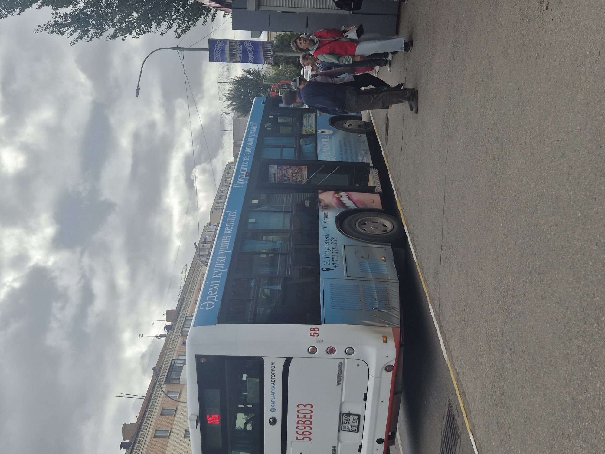 A bus with license plate 569BE03, manufactured by Yutong and part of the "Saryarka Avtoprom" company, is parked at a bus stop on a cloudy day. The bus displays the number 16 in red on its front and 58 on its side. The bus is blue and white with advertisements. On the side of the bus, there is text in Cyrillic which translates to "Beautiful smiles are coming!" and "Come for a healthy smile!". Also visible on the bus is a phone number: +7 771 278 12 28. A group of people are standing on the sidewalk near the bus, some appearing to be waiting. In the background, there are buildings and a street lamp. The scene is likely in Kokshetau, Kazakhstan, given the Cyrillic script and the known presence of "Saryarka Avtoprom" buses there.