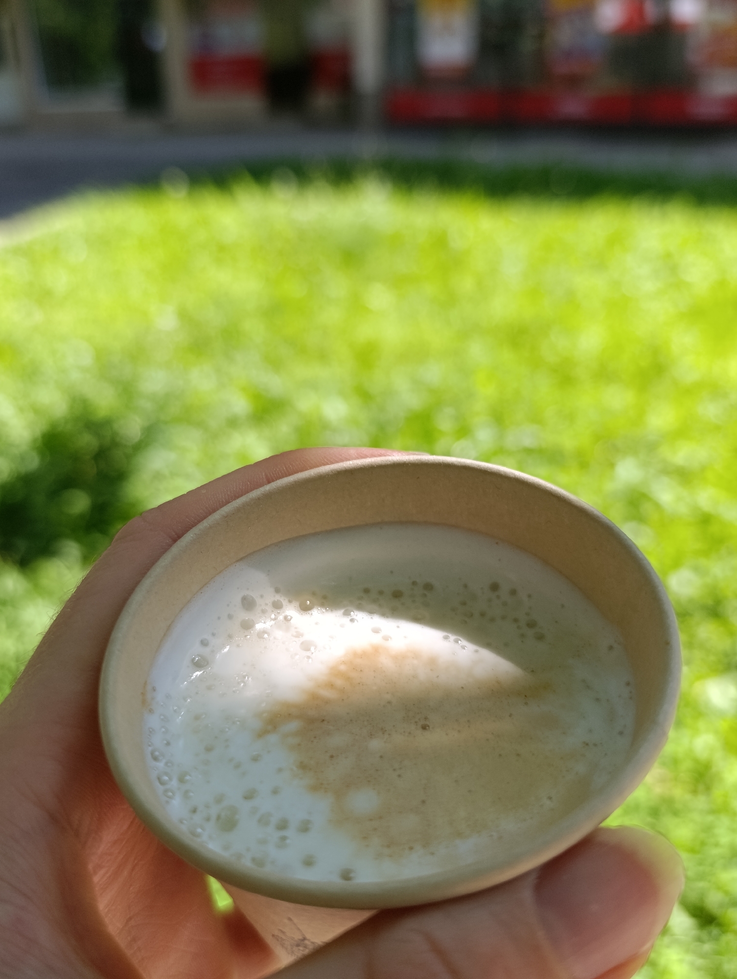 A hand holds a disposable coffee cup filled with a frothy beverage, possibly a latte or cappuccino, against a backdrop of blurred green grass and a suggestion of buildings. The image is taken outdoors, likely on a sunny day given the lighting and shadows. The focus is on the coffee cup and the hand, with the environment softly out of focus.