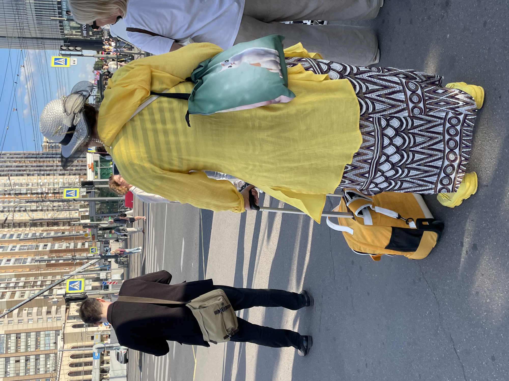 This is a medium shot, full-body photograph of several individuals at a crosswalk in Saint Petersburg, Russia, on a sunny day. The foreground features a woman dressed in a bright yellow, loosely draped tunic over a patterned skirt with a geometric design. She wears bright yellow shoes and a wide-brimmed, light-colored hat adorned with a black ribbon and decorative elements. A green tote bag with a cartoonish white bunny illustration hangs from her left shoulder. She is holding the handle of a yellow rolling suitcase.
To the left of the woman, a man in a black suit and black pants is walking away from the camera. He carries a tan messenger bag over his right shoulder. His black leather shoes are visible.
In the background, several other people are crossing the street. Pedestrian crossing signs with a blue background and white triangle depicting a person are visible on poles, indicating a regulated crosswalk. Tall, modern apartment buildings with numerous windows dominate the cityscape, with overhead power lines and traffic lights also present. The street is paved with asphalt, and shadows are cast on the ground, indicating the sun's position. The overall impression is a candid street scene in a bustling urban environment.