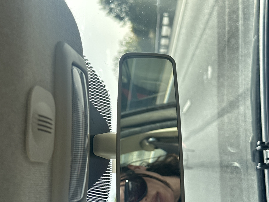 The image depicts an interior view from a moving vehicle, looking forward through a windshield. Centrally positioned is a rectangular rearview mirror, reflecting the vehicle's driver and interior elements. The driver, visible only as the lower left portion of their face, wears dark-framed sunglasses, has dark hair, and appears to have slightly parted lips and a silver hoop earring in their left ear. Above the rearview mirror, a light-colored overhead console houses a clear, ribbed cabin light lens and an oval-shaped grille with vertical slits. The windshield itself shows some smudges or dust, particularly in the lower section, and a black dotted frit band along its top edge.
Through the windshield and reflected in the mirror, a multi-lane highway or motorway is visible, featuring grey asphalt and clear white lane markings. To the right of the roadway, a light-colored barrier or railing with vertical supports, possibly part of a bridge or overpass, is present. Beyond this structure, green trees and foliage line the route under a bright, possibly hazy, sky. The vehicle is in motion on this highway.
The scene's environment, a multi-lane highway flanked by vegetation and a bridge structure, is consistent with major road networks such as the M2 or A2 motorways that traverse the Borough of Swale, United Kingdom. However, no specific identifying landmarks are present within the image to pinpoint an exact location within the borough.