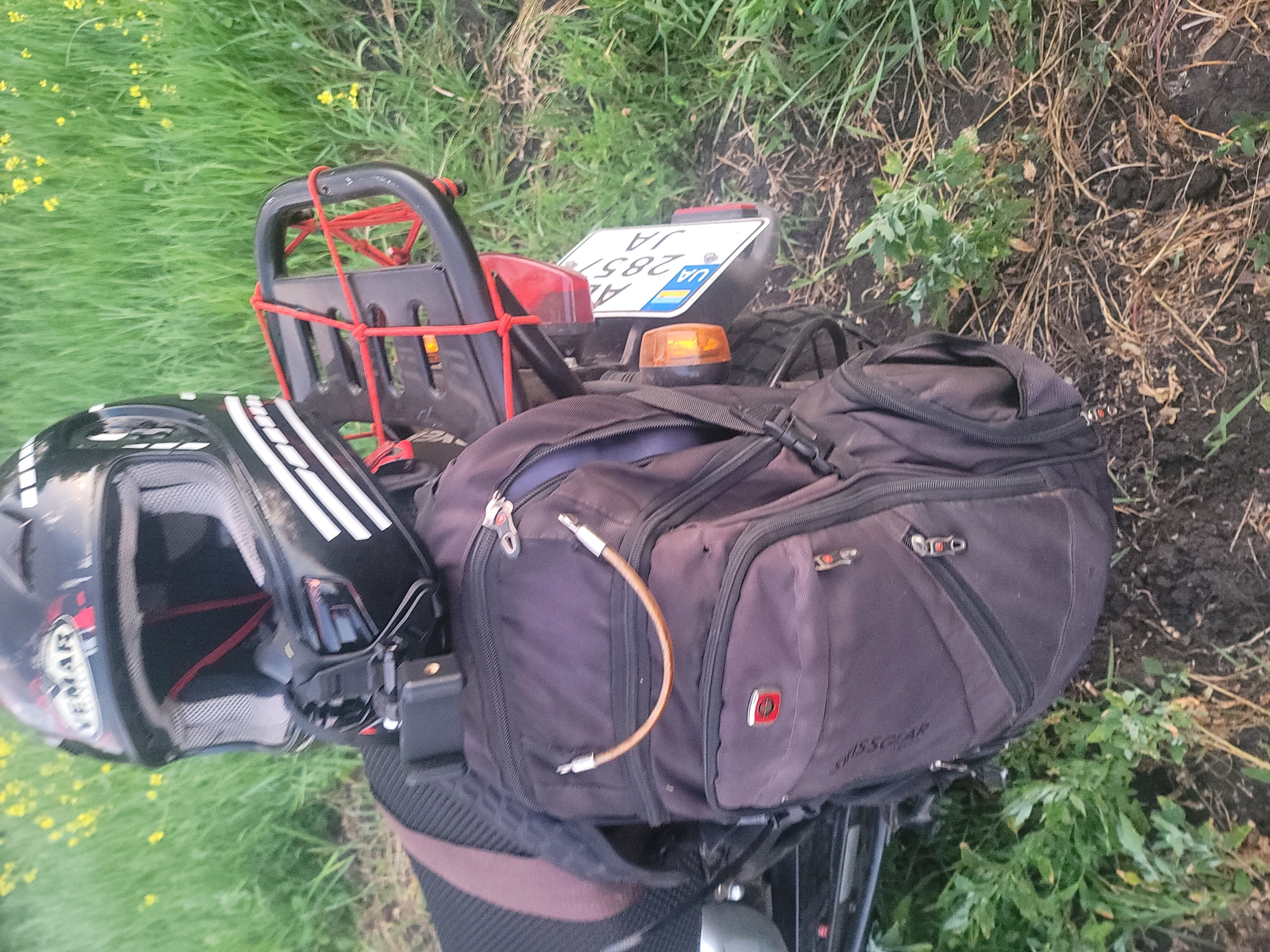 An over-the-shoulder, medium shot captures a black and white motorcycle helmet, a black backpack, and a license plate, all resting on the seat of a motorcycle that is parked on uneven ground amidst tall green grass and some yellow wildflowers. The motorcycle is positioned at a slight angle to the right. The helmet, with its visor down, is secured to the backpack with a black strap. A small action camera is mounted on the front of the helmet. The backpack is dark purple with a brown strap extending from it. A U-shaped brown cord is attached to the front of the backpack. The license plate, which is white with blue and yellow markings and black text, is visible on the back of the motorcycle. The motorcycle is not entirely visible, but a portion of its rear fender and tail light are discernable. The background is dominated by dense green grass.