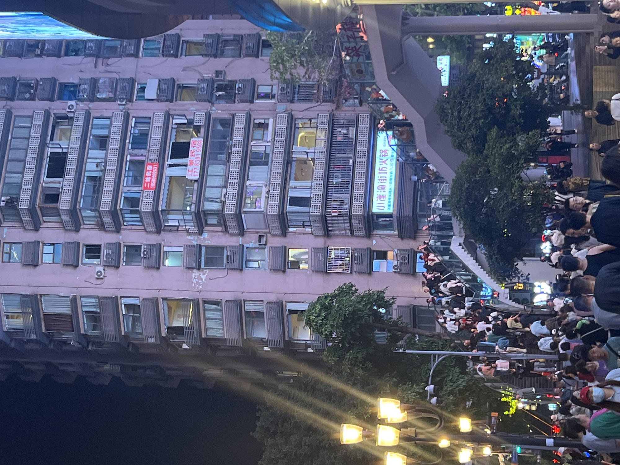A medium shot captures a street scene in Yuzhong District, Chongqing, China. A tall, densely windowed building stands to the left, with air conditioning units installed in many windows. Several banners or signs are hung on the building's facade. To the right, a crowded pedestrian walkway teems with people, mostly facing the camera. Streetlights provide illumination in the foreground, and trees line the street. The overall environment suggests an urban area during nighttime.