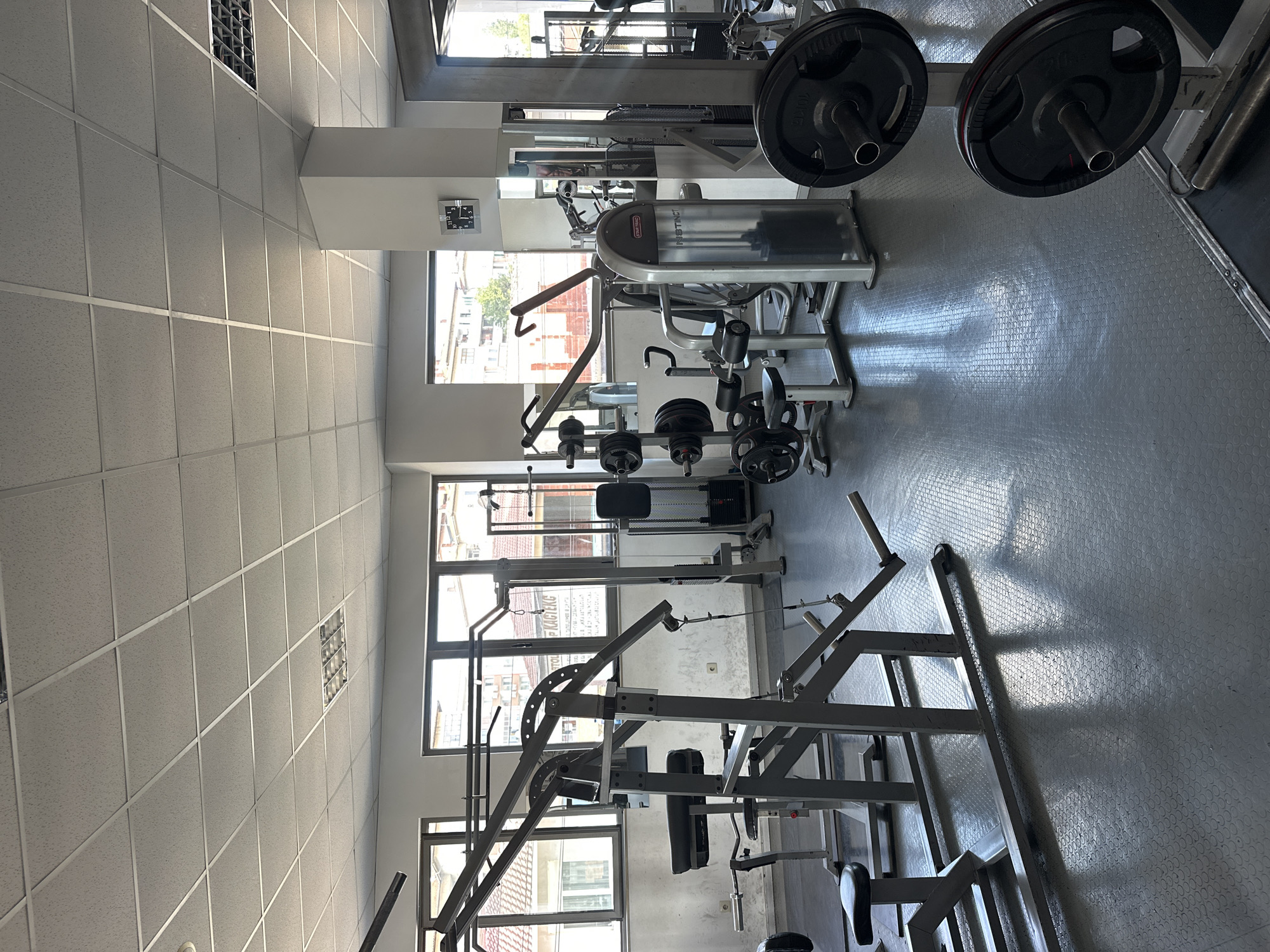 The image presents the interior of an empty gym, illuminated by abundant natural light entering through large windows. The setting appears to be a commercial fitness facility, devoid of people or ongoing activities.
The gym features a white grid-patterned drop ceiling with acoustic tiles and integrated lighting fixtures. The walls are light-colored, and the floor is covered with grey, textured rubberized mats, characteristic of gym flooring. Sunlight streams through the windows, casting prominent shadows of the equipment and window frames across the floor, indicating a bright, sunny daytime.
Various exercise machines and free weights are visible throughout the space. Notable equipment includes a multi-station weight machine prominently displaying "STARTRAC" and "INSTINCT" branding. Nearby, weight plates marked "10 KG" and "20 KG" are stored on a rack. Other machines, including what appears to be a lat pulldown station and possibly a leg press, are also present.
A black square wall clock with white numerals and hands is mounted on a white pillar between two window sections. Through these windows, glimpses of surrounding residential or commercial buildings are visible, suggesting an urban or suburban location, consistent with Veliko Tarnovo, Bulgaria. Some small, illegible text is present on one of the windowpanes.