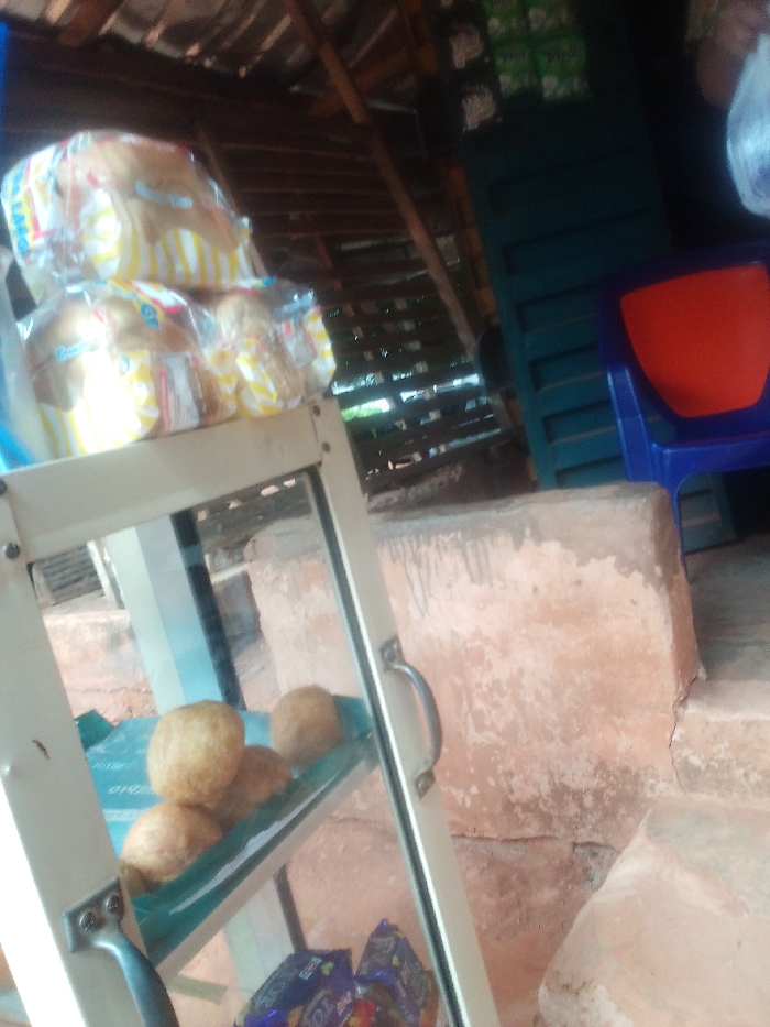 A small, open-air food stall in Auchi, Nigeria, is located at coordinates 7.063 latitude and 6.274 longitude. Three loaves of bread, wrapped in clear plastic with yellow and white stripes, are stacked on top of a white metal stand with glass shelves. The stand is situated in front of a concrete wall and a blue plastic chair with an orange seat. Four round, brown pastries sit on the bottom shelf of the stand, covered in green plastic wrap. Chocolate candies and a single, green plastic bag are visible on the bottom shelf as well. There are no people visible in the image. The overhead structure is made of wood and appears to be a roof. A wooden, slatted wall with a door is visible to the right. The floor appears to be made of concrete and is uneven. The area is dusty, and the concrete wall is covered with dirt and water stains. The scene is most likely a street vendor's stall, located on a busy street. The image is taken from a low angle.