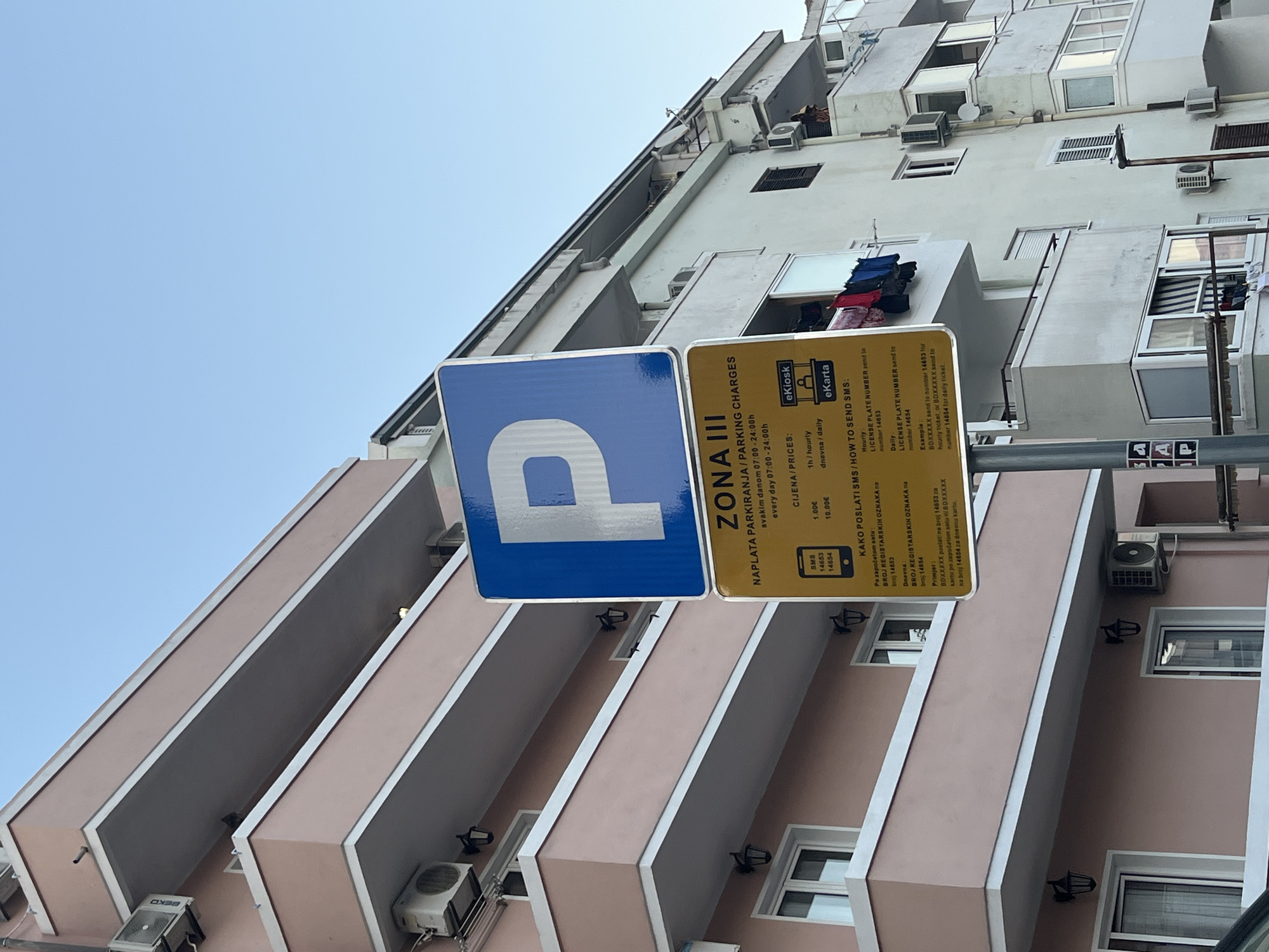 A close-up, eye-level, outdoor shot captures a blue and white parking sign with a large "P" symbol and a yellow information sign below it, set against a clear blue sky and the facade of a light pink apartment building with multiple balconies. The pink building features white trim around its balconies and air conditioning units mounted on its exterior. To the right of the pink building, a portion of a lighter-colored apartment building is visible, also with balconies.
The yellow sign provides parking information, with text in what appears to be Croatian. It indicates "ZONA III" (Zone III) and details "NAPLATA PARKIRANJA / PARKING CHARGES" valid from "07:00 - 24:00h every day." It also lists "CIJENA / PRICES," with "1h / hourly" costing "1.00€" and "dnevna / daily" costing "10.00€." Further down, under "KAKO POSLATI SMS / HOW TO SEND SMS," it instructs users to send their license plate number to specific numbers for hourly and daily tickets, providing an example. Icons for "eKiosk" and "eKarta" are also present.
A metallic pole supports the two signs. Below the yellow sign, on the pole, are other smaller signs with black and white symbols. The building on the right has windows, balconies, and air conditioning units. Laundry is hanging on a clothesline on one of the balconies of the building on the right. A person, possibly a woman wearing a dark garment, is visible standing on a balcony of the building on the right.