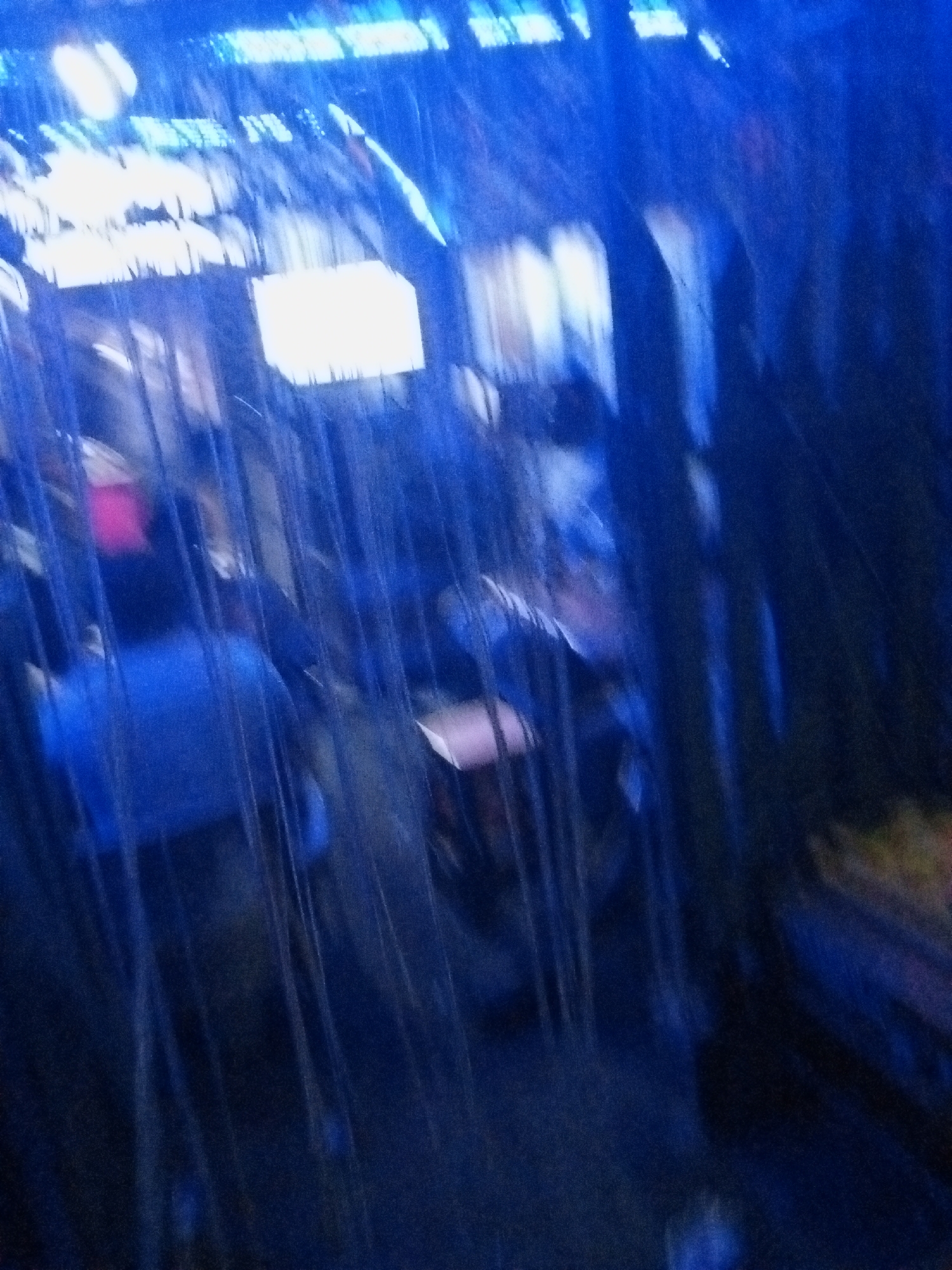 The image appears to be taken indoors in a dimly lit area, possibly inside a building with a partially exposed or unfinished ceiling structure. The scene is dominated by a blue hue, with vertical lines or structures (possibly bars or posts) in the foreground creating a sense of enclosure. Several blurry figures are discernible in the background, one with a red object near their head. There are other objects and surfaces within the room, but they are indistinct. The location, Ado, Nigeria, is not discernable from the image.