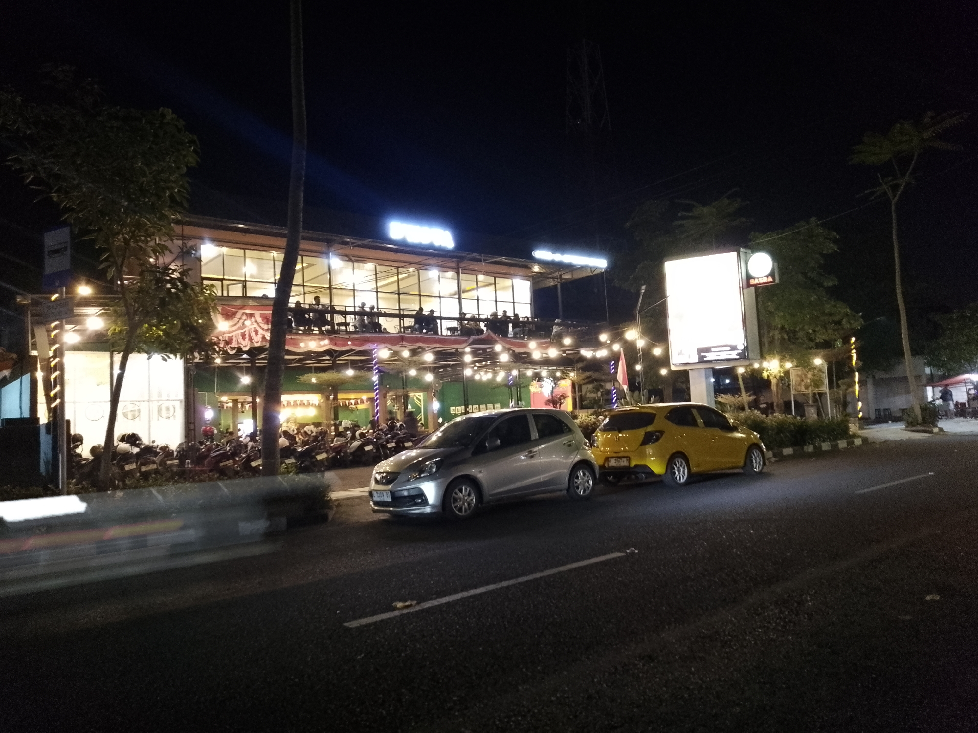 The image captures a nighttime scene in Lamongan, Indonesia, focusing on a brightly lit establishment. The building, illuminated by internal lights and numerous string lights, appears to be a restaurant or cafe with a second-floor outdoor seating area. Several people are visible seated at tables on the upper level. In front of the building, on the ground level, a row of parked motorcycles is visible.
On the street in front of the establishment, two cars are parked: a silver hatchback on the left and a yellow hatchback on the right. The silver car has its headlights on, and the yellow car has its license plate partially visible. The street is paved and marked with white lines. The overall atmosphere is that of a busy commercial area at night.