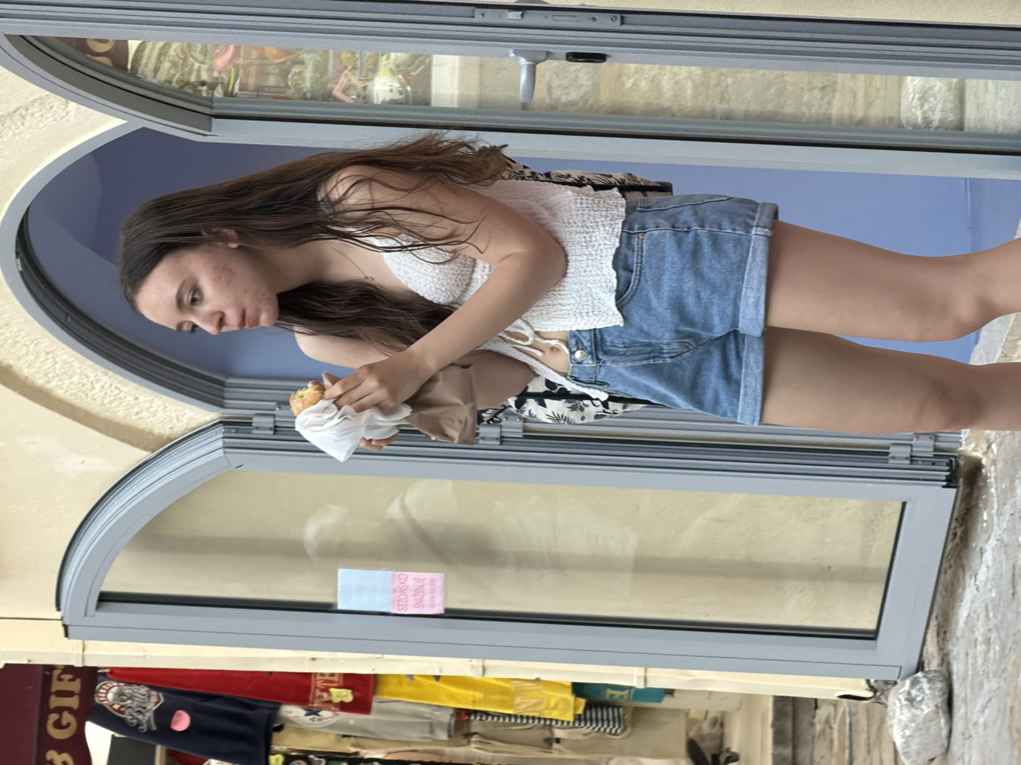 A young woman with long, dark brown hair is standing outside a shop in Budva, Montenegro, holding and eating a pastry wrapped in white paper. She is wearing a white crocheted tank top and denim shorts. The shop has large glass doors with arched frames, and the building exterior appears to be made of light-colored stone. Behind her, through the glass, are reflections and some items for sale. To her left, on the exterior wall of the shop, are two signs, one with text that appears to be in a Slavic language, reading "SEZONSKO SNIŽENJE" which translates to "Seasonal Discount". The ground in front of the shop is paved with stone, and a large light-colored rock is visible on the left side of the frame. The scene appears to be a street or market area in a European city.