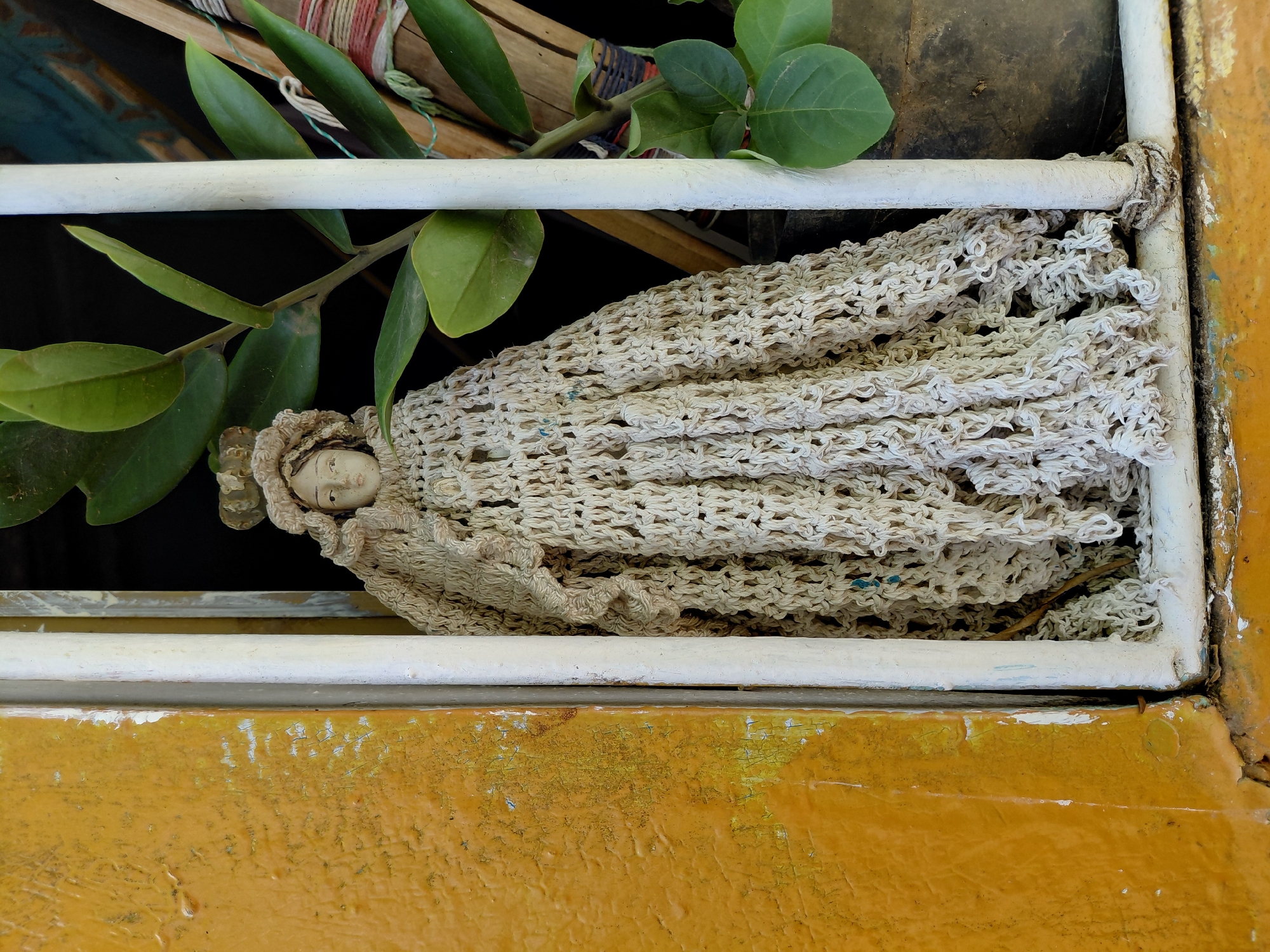 A doll with a crocheted dress and headdress is positioned behind a white barred window. The doll's face is visible, with painted features. The window frame and bars are white, and a yellow, peeling painted wall is to the left of the window. A green leafy plant is visible to the right of the doll, and behind it, a dark pot and some wooden objects are present. The scene appears to be outdoors, possibly on a street in Santa Ana, Venezuela.