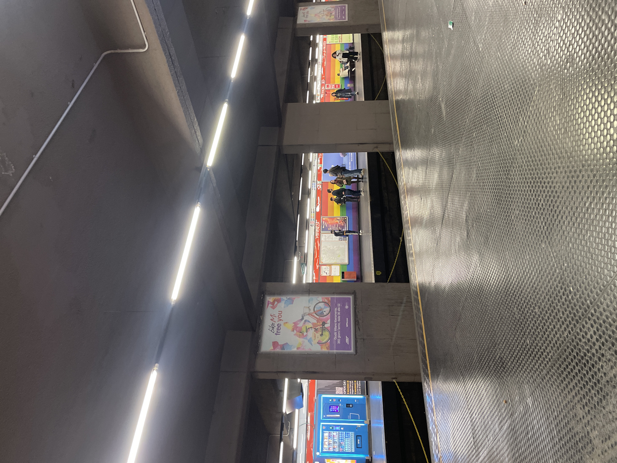 The image shows the platform area of a Milan Metro station. The lighting consists of fluorescent tubes mounted on the ceiling. Advertisements and vending machines are visible on the platform level. Several people are waiting on the platform, with rainbow-themed murals on the wall behind them. A sign on a pillar shows an advertisement for "BikeMi Free You". The floor is textured metal, and the area has a utilitarian, transit-oriented aesthetic.