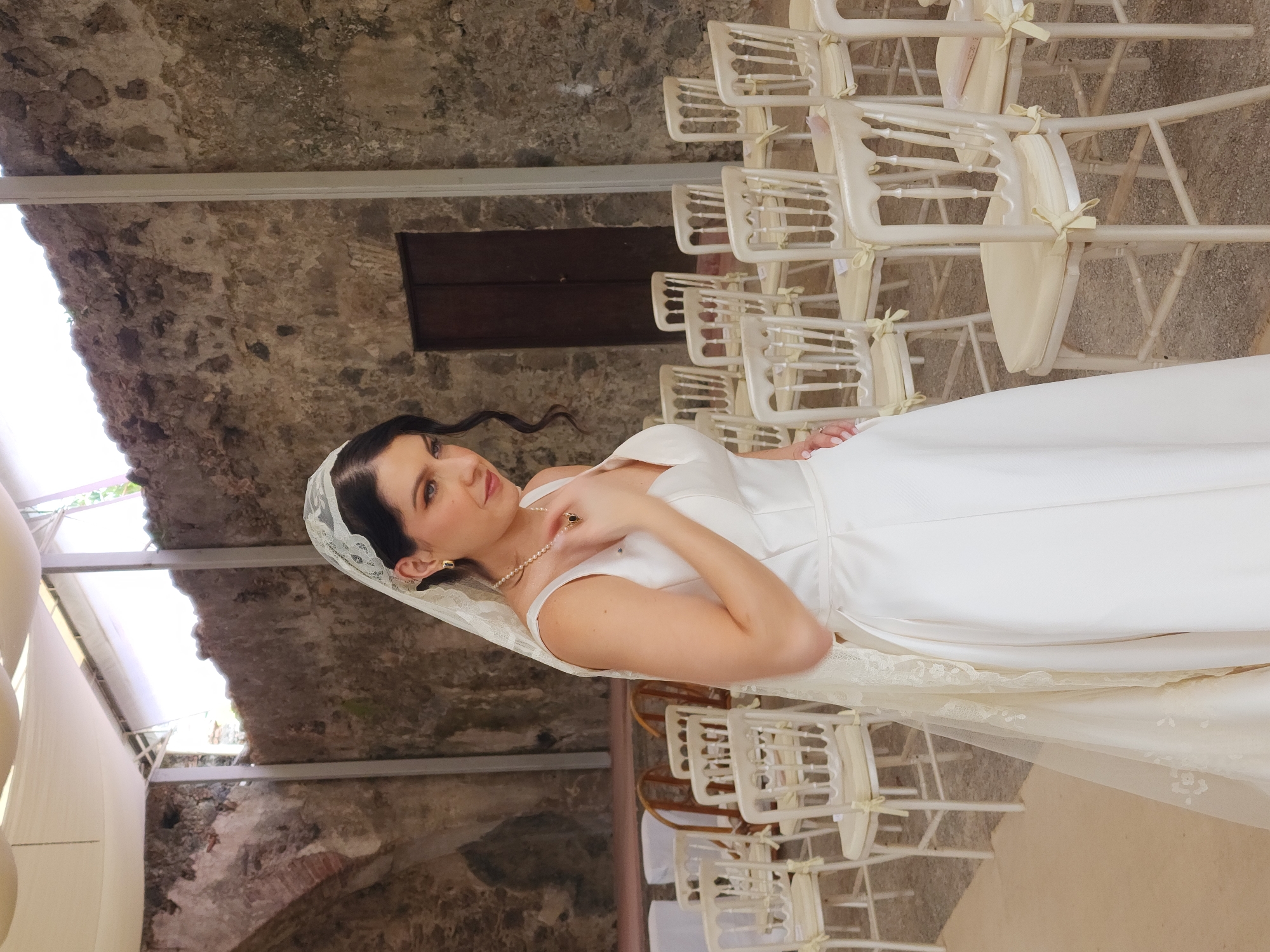 A vertical, medium shot features a woman in a white wedding gown, positioned in front of rows of empty white chairs. She is adorned with a pearl necklace, lace veil, and gold earrings. Her left hand is raised and touches the jewelry on her chest. The setting appears to be an outdoor event space with a stone wall, a covered ceiling, and a small wooden door on the stone wall. The location is Unknown, Unknown.
