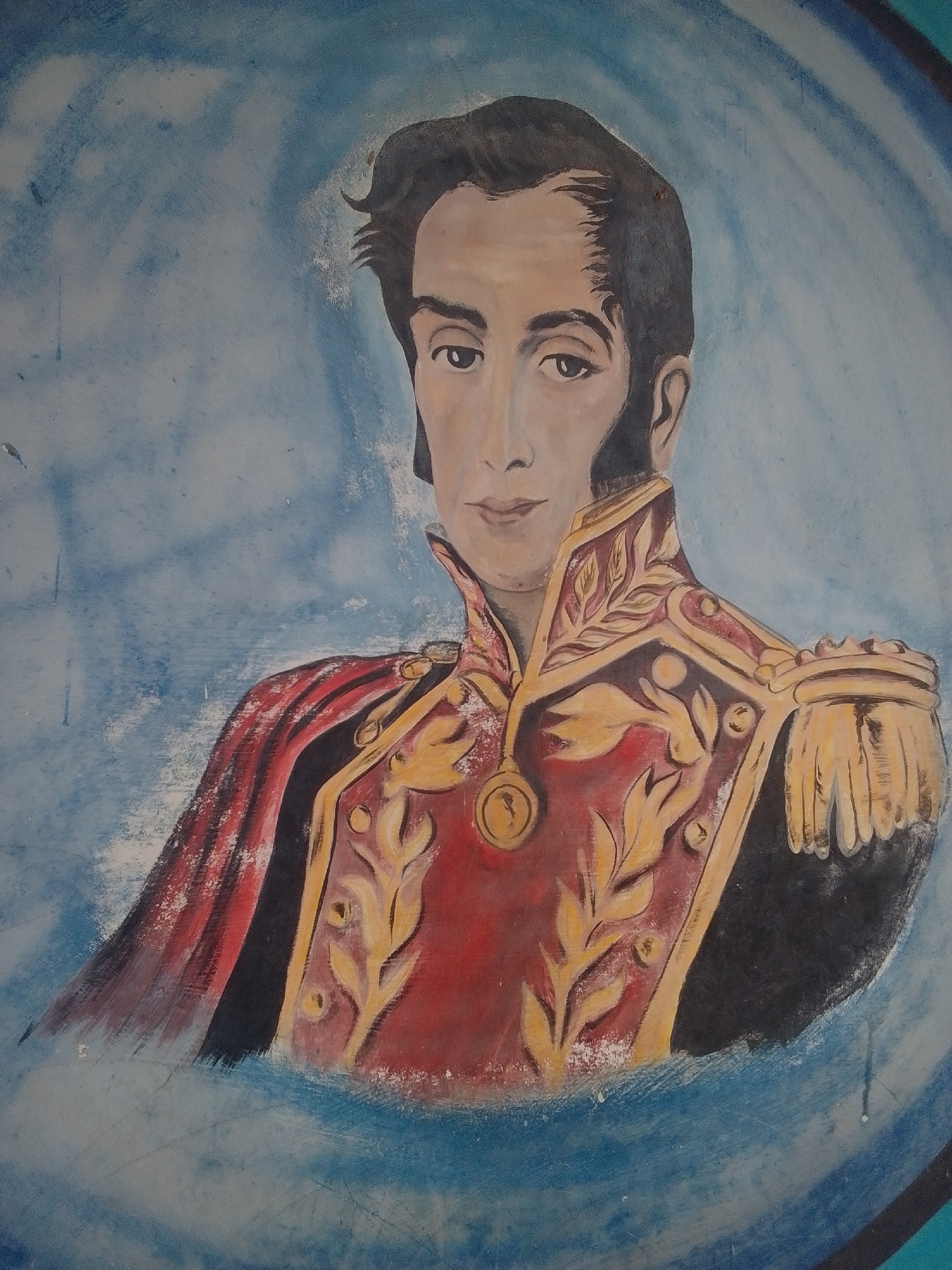 This is a painting on a wall depicting a portrait of a man in a military uniform. The man has dark hair styled with a side part and prominent sideburns. He has a fair complexion, dark eyes, and a serious expression. He is wearing a red jacket adorned with intricate gold detailing on the collar, shoulders, and front. The jacket also features a red cape draped over his left shoulder, lined with black. A circular medallion hangs from a chain around his neck. The background is a textured blue, suggesting a sky or water. The style of the painting is somewhat stylized and appears to be a mural, possibly on an exterior wall given the textured appearance and some visible paint imperfections. The location context provided is La Tagua, Venezuela, which might suggest the subject is a historical figure important to Venezuelan history. There is no visible text in the image.