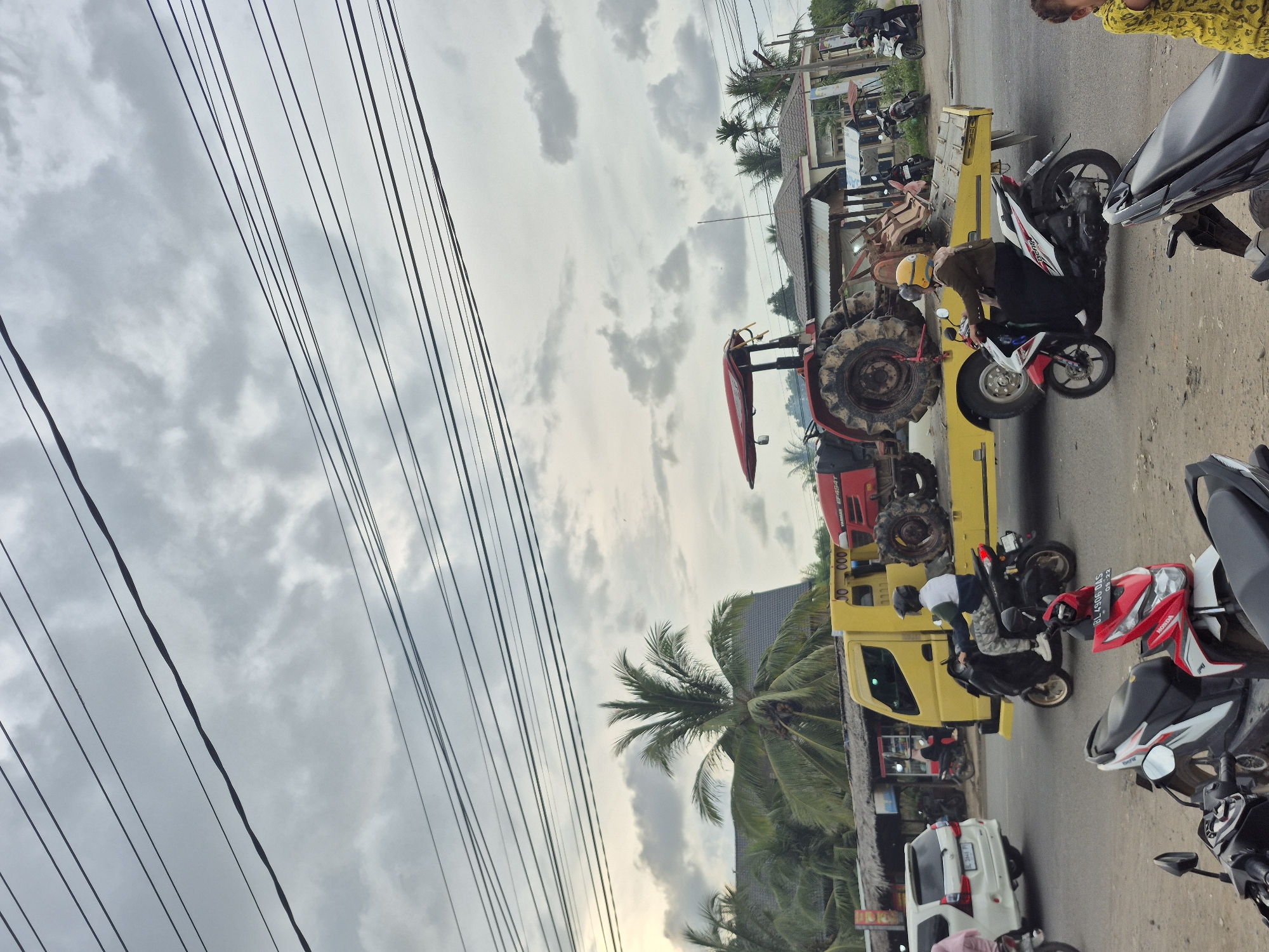 A red tractor is transported on a yellow flatbed truck in Tingkeummanyang, Indonesia. The roadside scene shows an overcast sky, overhead power lines, and numerous parked motorcycles. Nearby are buildings with thatched roofs and palm trees. People are on the road.