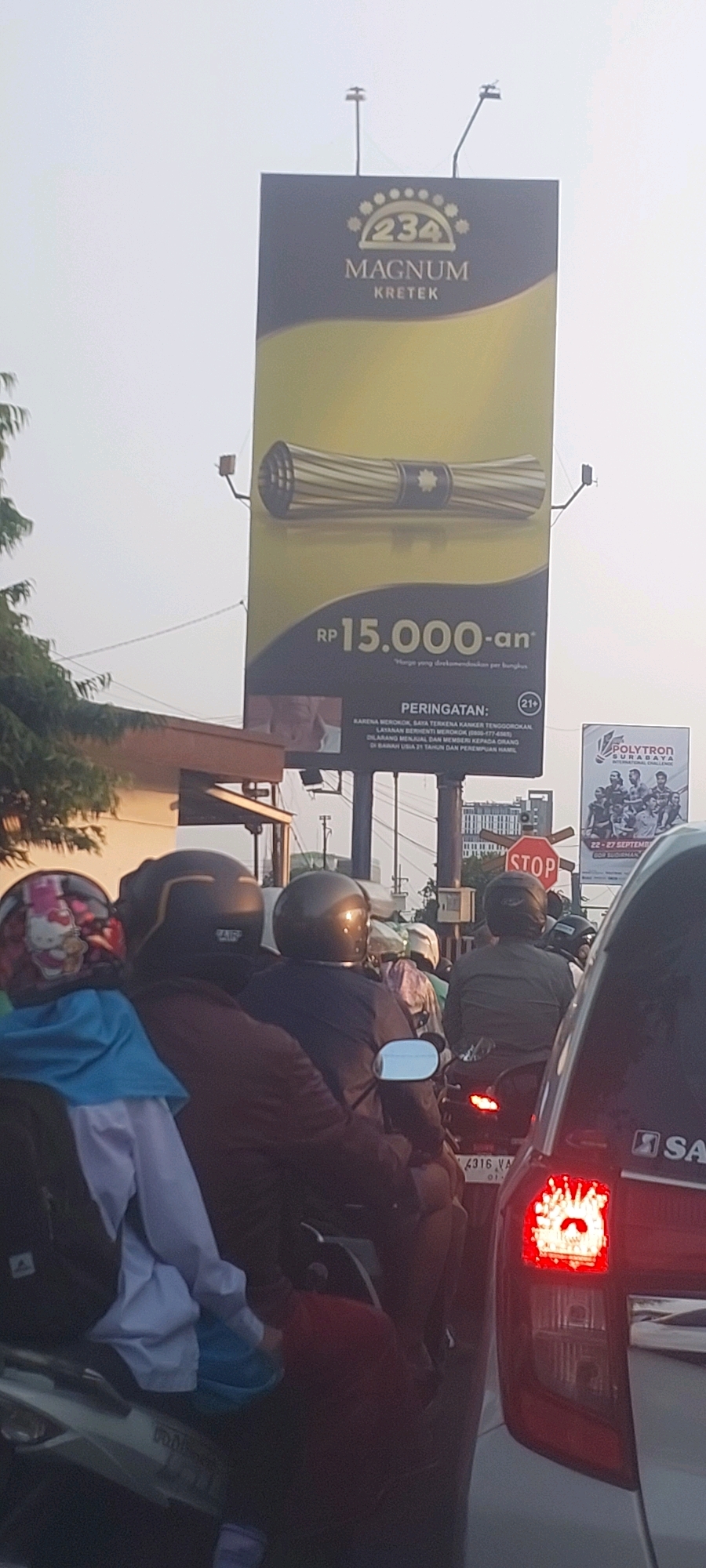 The image captures a street scene, likely in Surabaya, Indonesia, where a traffic jam is occurring. In the foreground, several motorcycles are stopped. On the first motorcycle from the left, a person wearing a helmet with a Hello Kitty design is seated. Behind them, two individuals are on another motorcycle, both wearing dark helmets. The second person from the left appears to be wearing a red shirt. A dark-colored car's taillight is visible on the far right, indicating its presence in the traffic.
Dominating the upper portion of the image is a large billboard advertising "234 Magnum Kretek". The billboard displays the product name, the price "RP 15.000-an", and a warning in Indonesian: "PERINGATAN: KARENA MEROKOK, SAYA TERKENA KANKER TENGGOROKAN. LAYANAN BERHENTI MEROKOK (0806-177-4585). DILARANG MENJUAL DAN MEMBERI KEPADA ORANG DI BAWAH USIA 21 TAHUN DAN PEREMPUAN HAMIL." This translates to: "WARNING: BECAUSE OF SMOKING, I HAVE THROAT CANCER. QUIT SMOKING SERVICE (0806-177-4585). IT IS FORBIDDEN TO SELL AND GIVE TO PERSONS UNDER 21 YEARS OF AGE AND PREGNANT WOMEN." A small circular icon indicating "21+" is also present.
To the right of the main billboard, another smaller billboard is visible, promoting "POLYTRON SURABAYA INTERNATIONAL CHALLENGE" with dates "22-27 SEPTEMBER". A traffic stop sign is also visible, positioned above the motorcycles. The environment appears to be a bright, possibly late afternoon or early morning, with a bright sky and some trees lining the street on the left. The presence of multiple vehicles and a billboard suggests an urban setting.