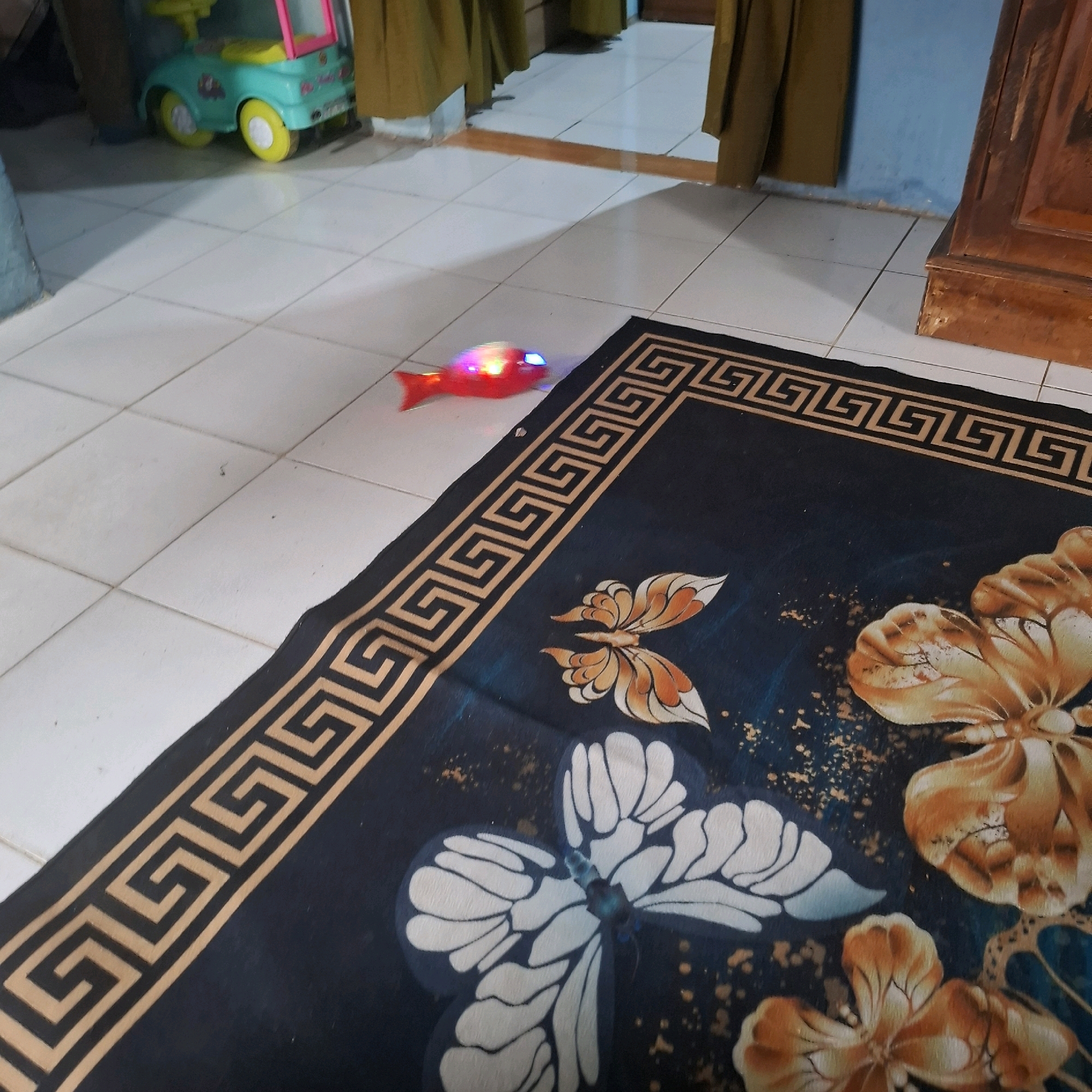 In a room with white tiled flooring, a red toy fish with flashing lights is moving across the floor. A black rug with a geometric border and a butterfly design is laid out in the foreground. In the background, a green and yellow toy car is parked near a doorway. The scene appears to be indoors, likely a home, in Bandar Lampung, Indonesia.