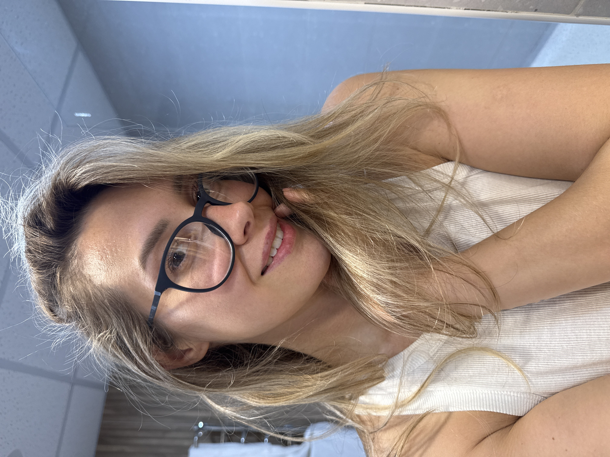 A medium close-up shot shows a young Caucasian woman with blonde hair and glasses, smiling softly while looking towards the viewer. She is wearing a white ribbed tank top. Her left hand is brought up to her cheek and chin, obscuring part of her face. The background appears to be a bathroom with a light blue tiled wall and a mirrored surface reflecting a ceiling light. The image does not provide any specific visual cues to indicate its location in Nebug, Russia.