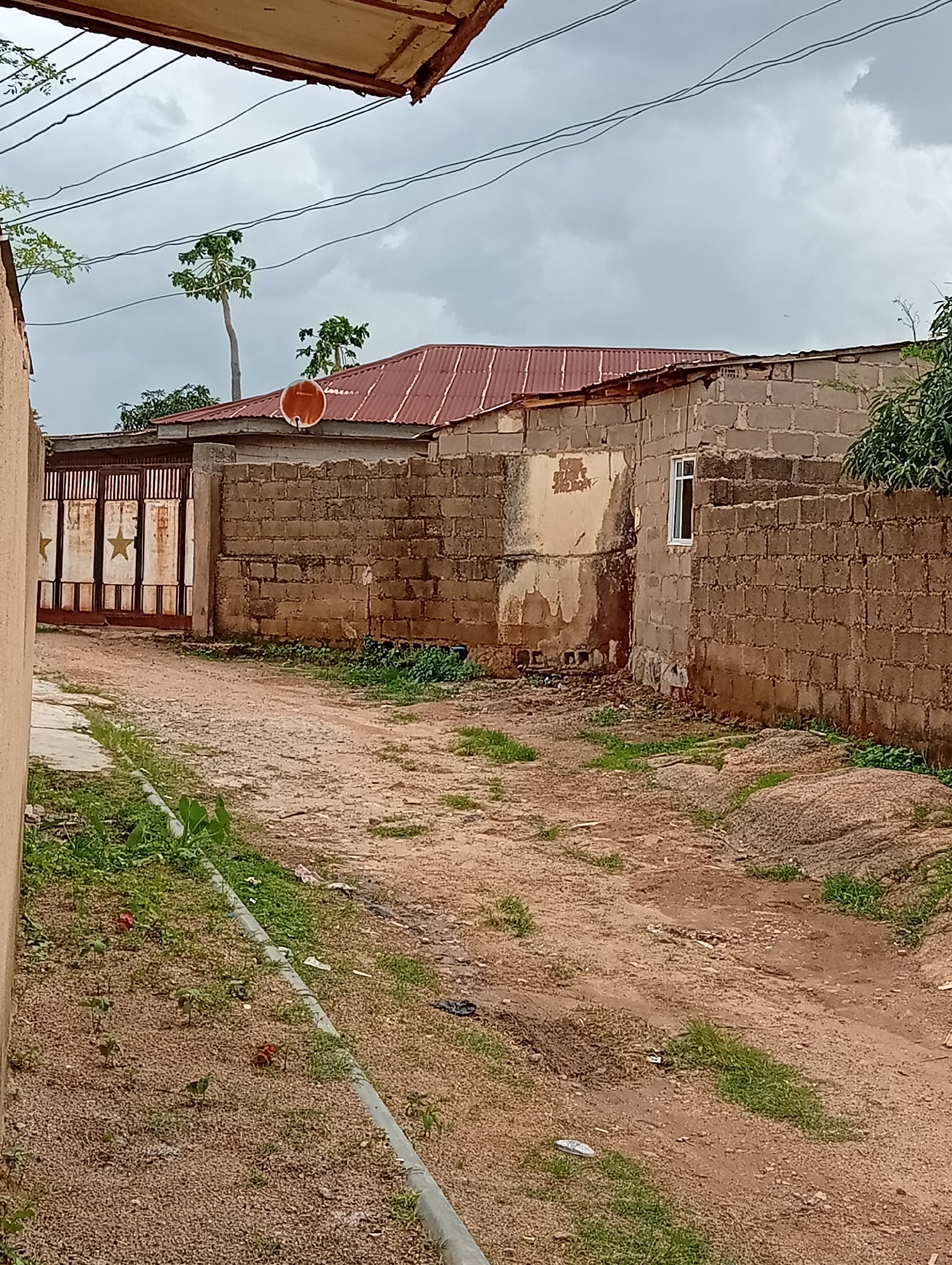 The image depicts a dirt road leading towards a large, ornate metal gate with star designs. Beyond the gate, a fence made of concrete blocks encloses a property. A house with a corrugated metal roof stands to the right of the gate, also constructed from concrete blocks. The house has a small, white-framed window. A satellite dish is mounted on the roof. The area appears to be somewhat overgrown with grass and vegetation lining the sides of the road. The sky is overcast, suggesting a cloudy or possibly rainy day. There are power lines stretching across the upper part of the image. The overall scene is an outdoor view of a residential area, possibly in a developing or rural setting. There is no visible text in the image. The time of day is unclear due to the overcast conditions. The location context is Jos, Nigeria.