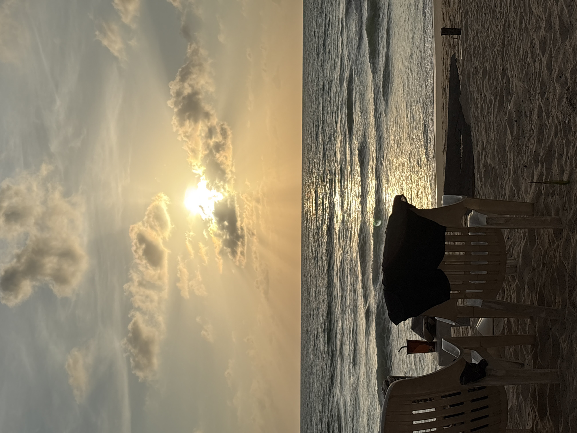 The image depicts a serene beach scene at sunset or sunrise, likely in Choeng Thale, Thailand. Two empty plastic beach chairs are positioned facing the ocean, with a black garment draped over the back of the chair on the right. A beverage cup with a straw sits on a small table between the chairs. The sun is partially obscured by clouds, casting golden rays across the sky and shimmering on the water's surface. Gentle waves roll towards the sandy shore. The overall atmosphere is one of tranquility and relaxation.