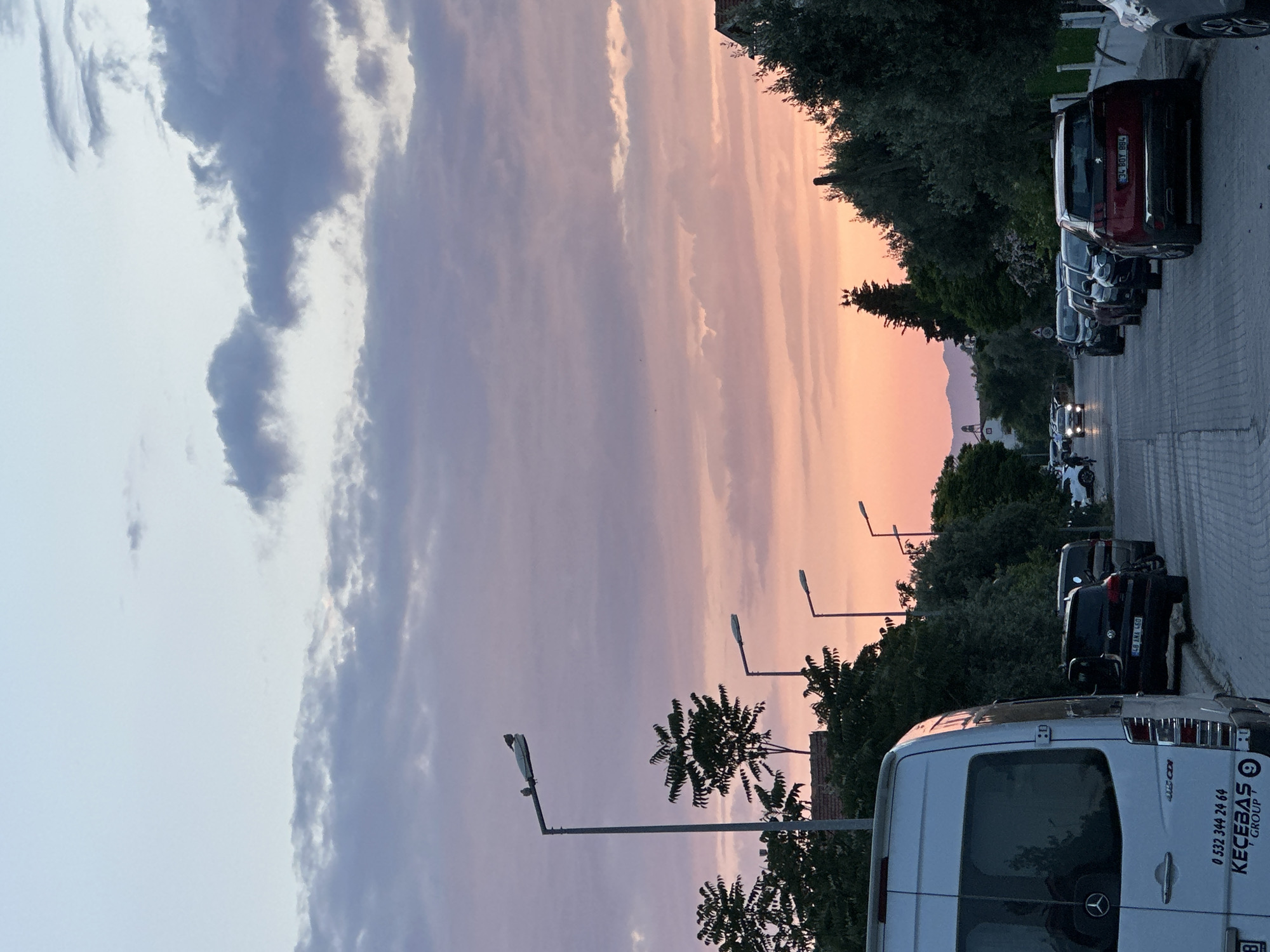 The image depicts a street scene in Fethiye, Turkey, under a sky transitioning between shades of blue, pink, and orange, likely during sunset. Along the right side of the frame, a row of parked vehicles, including a white van with "KEÇEBAS GROUP" branding and phone number ("0 532 344 24 64") on the rear, are lined up along a paved road. The license plates of some vehicles, like "34 BOY 884" on a red car, are visible. Streetlights punctuate the tree line separating the road from the sky. In the background, a suggestion of mountains peeks through the gap created by the street, and the headlights of distant vehicles are noticeable. Trees, likely urban landscaping, line the side of the street. The street itself appears to be made of cobblestones.