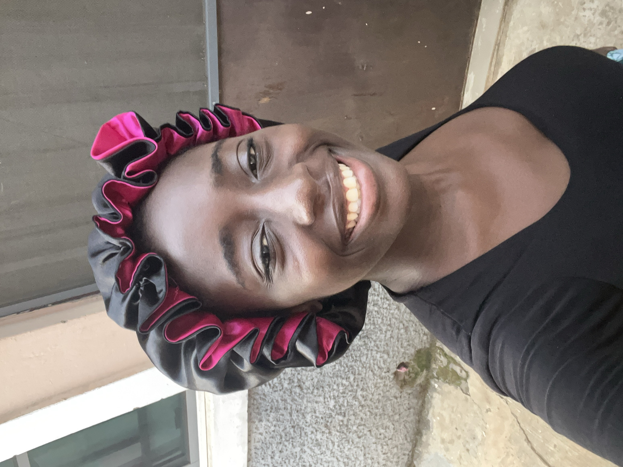 A woman with dark skin is smiling and looking at the camera. She is wearing a black shirt and a black and pink satin bonnet. The setting appears to be outdoors in Abuja, Nigeria, based on the architecture. The woman is standing on a concrete surface next to a building.