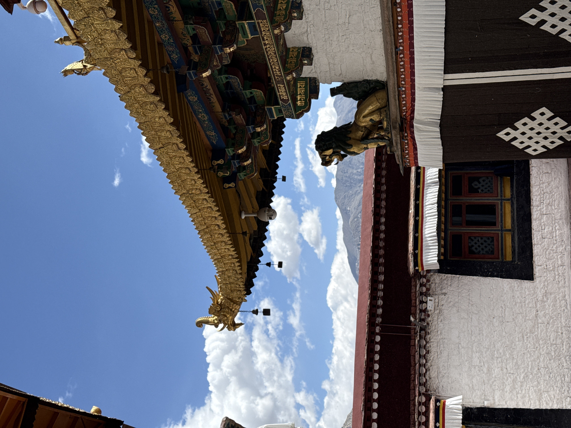 The image depicts a portion of a Tibetan-style building in the Chengguan District of Lhasa, China, with clear sky visible. The building features intricate gold trim along the eaves, a lion statue detail, and a window with ornate framework set against a white wall. The photograph is oriented at an unusual angle, showcasing both architectural details and the background sky.