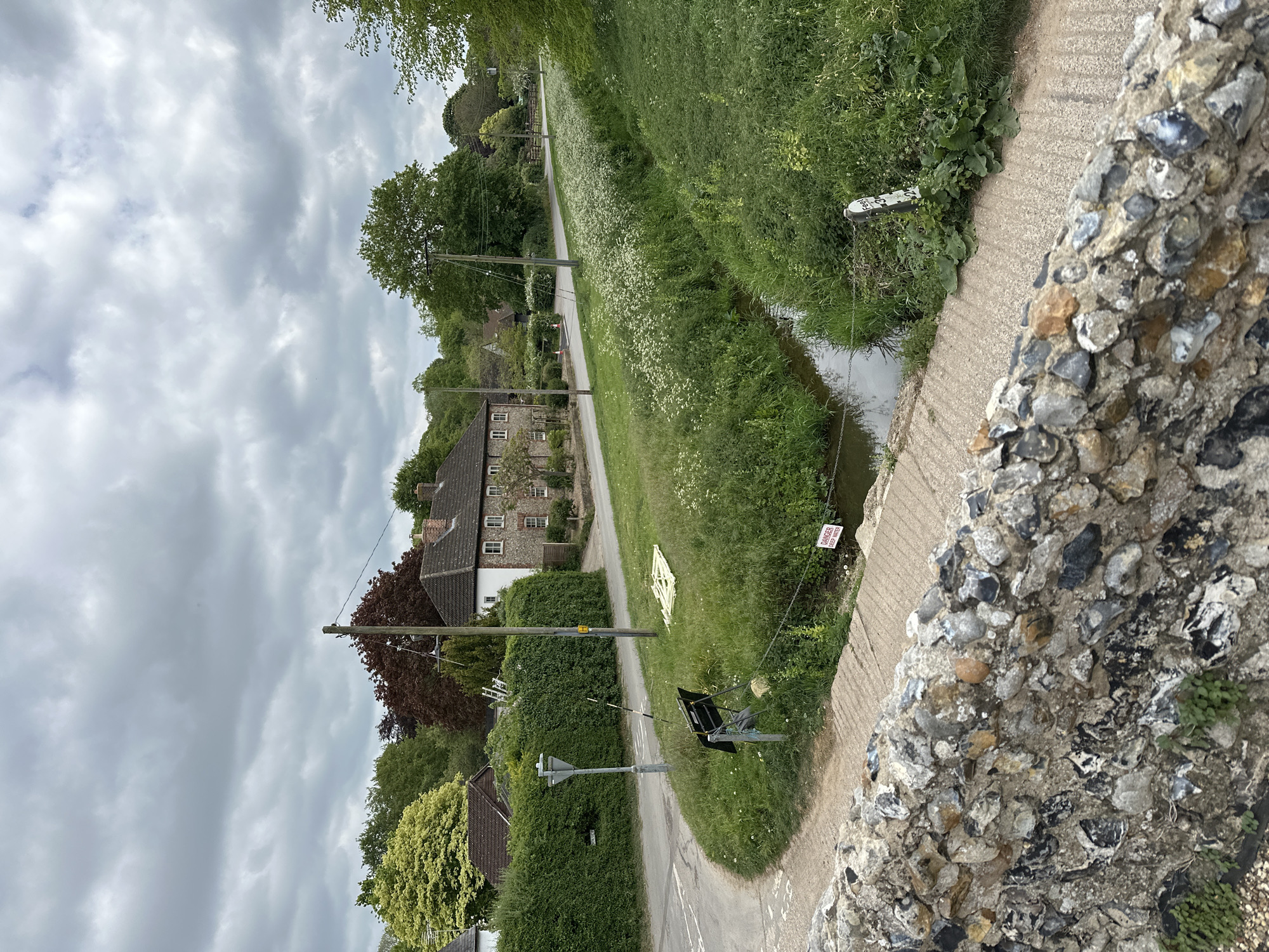 The image shows a view from a stone bridge in West Suffolk, United Kingdom. The view is primarily of a narrow country road lined with houses, a drainage ditch, and green vegetation. Along the road, there are utility poles with hanging wires. A damaged road sign lies on the grass next to the road. In the background is a building made of brick and stone, with a dark roof. The sky is overcast with dense clouds. The bridge's stonework is visible in the foreground, composed of various sizes and colors of stones embedded in mortar. A small warning sign is visible near the drainage ditch.