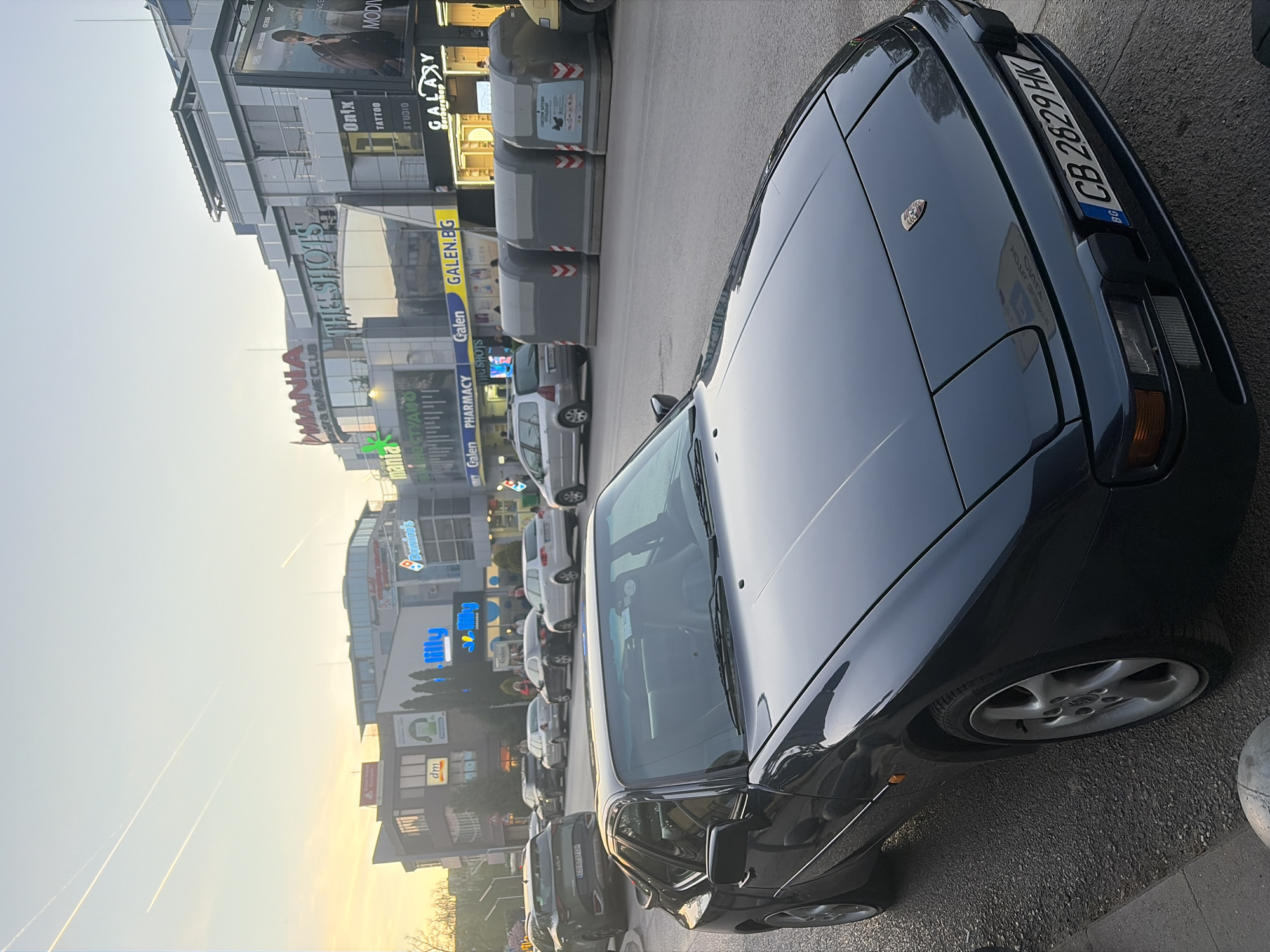 The image depicts a dark-colored Porsche 944 parked on a street in Sofia, Bulgaria. The car's license plate reads "CB 2829 HK" with the "BG" identifier. In the background, there is a busy street with several stores and restaurants, including Mania, Domino's, dm, lilly, Galen Pharmacy, Galaxy Barbershop, Onix Tattoo Studio, and a MODI store. Several other cars are parked or moving along the street. The sky is a light blue color with a hint of sunset, and there are visible contrails from airplanes in the distance. The scene appears to be a typical commercial area in Sofia.