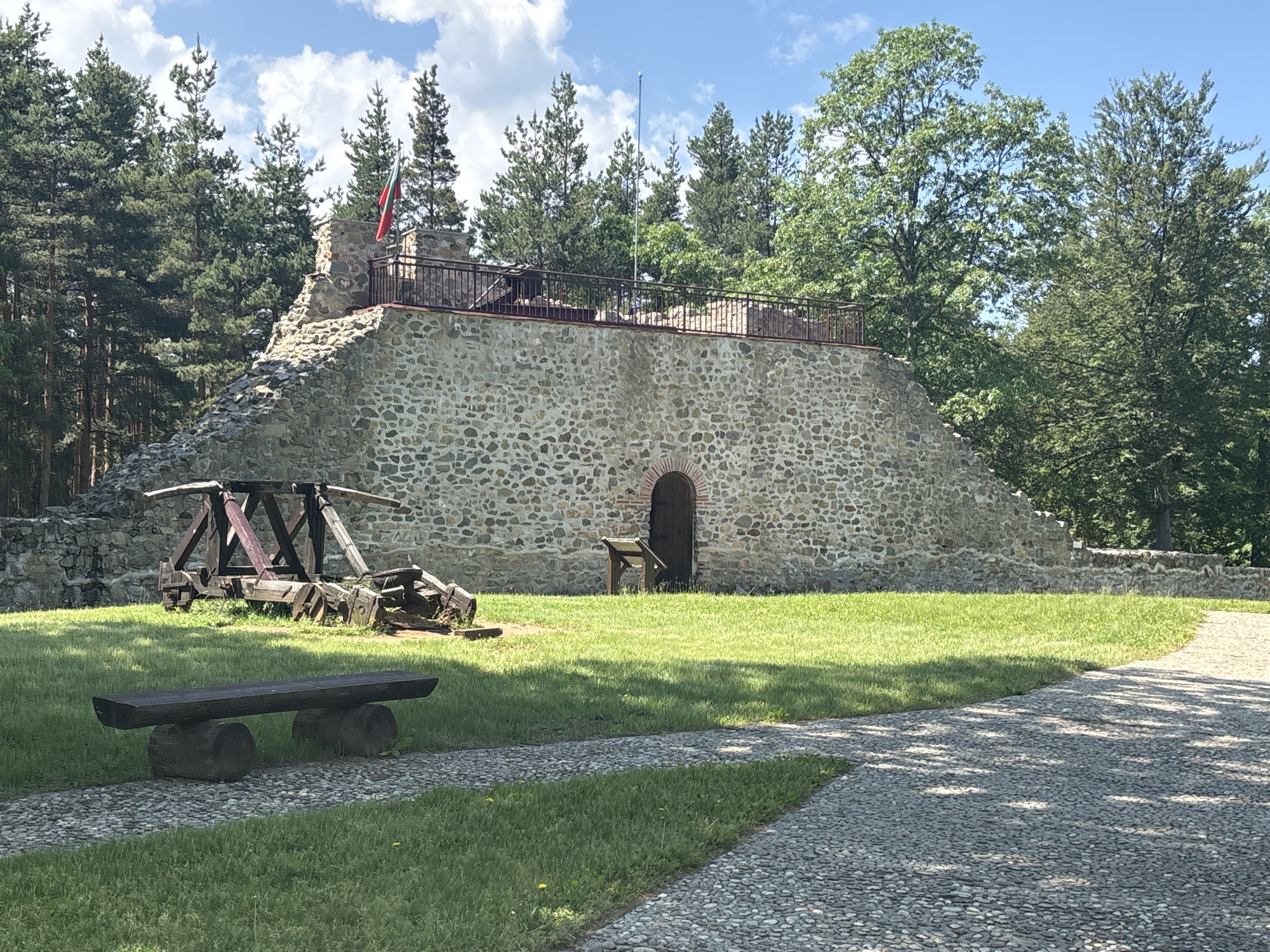 This detailed image depicts the ruins of Tsari Mali Grad Fortress located in Belchin, Bulgaria. The central feature is a substantial stone wall, constructed from irregular light-colored stones, exhibiting a weathered appearance. On the left, the wall ascends in a rough, sloped section, while the central and right portions feature a more vertical facade. A flat platform crowns the upper part of the wall, secured by a dark metal railing. A flagpole on the left side of this platform flies the Bulgarian flag, displaying horizontal white, green, and red stripes. Further along the platform to the right, a thin metallic pole, possibly a lightning rod, stands upright.
At the base of the central wall, a wooden door with a rounded archway is visible, next to which stands a small, dark wooden interpretive stand. Below the sloped section of the wall to the left, a large, dark wooden replica of a siege weapon, possibly a trebuchet or catapult, rests on the grass.
The foreground consists of a well-maintained green grass lawn, traversed by a winding path made of small, light-colored pebbles. A rustic wooden bench, fashioned from a dark timber plank supported by two large logs, is positioned on the grass to the left of the path. Portions of the path and grass are dappled with shadows from unseen foliage.
The background reveals a dense arrangement of tall coniferous trees, primarily pine, situated behind the left and central parts of the stone structure. To the right, tall deciduous trees with lighter green foliage are present. The sky above is clear blue with scattered white cumulus clouds. No people or animals are visible in the scene.